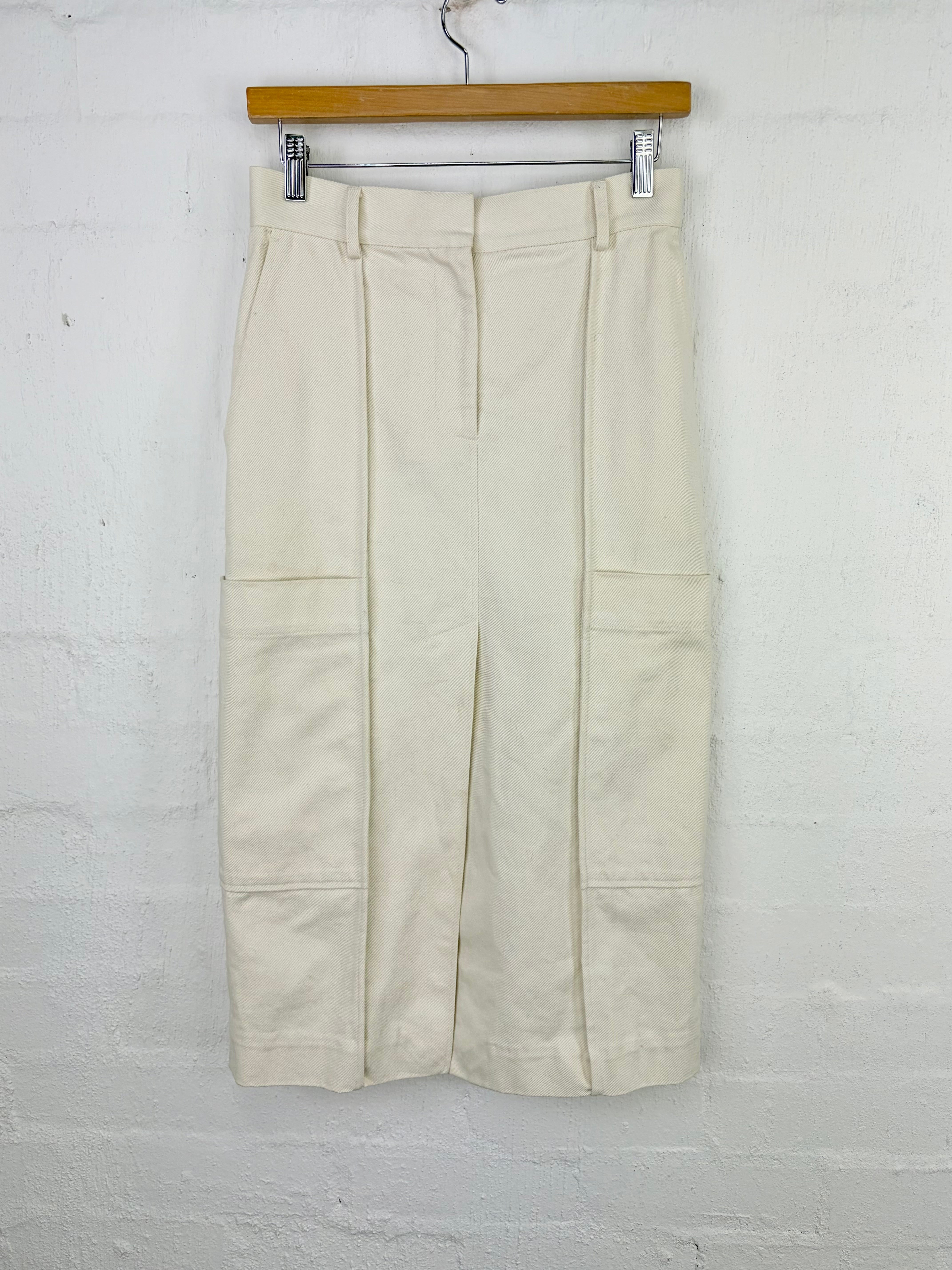 A pair of cream-colored pleated cargo-style pants hanging from a wooden hanger against a white painted brick wall.