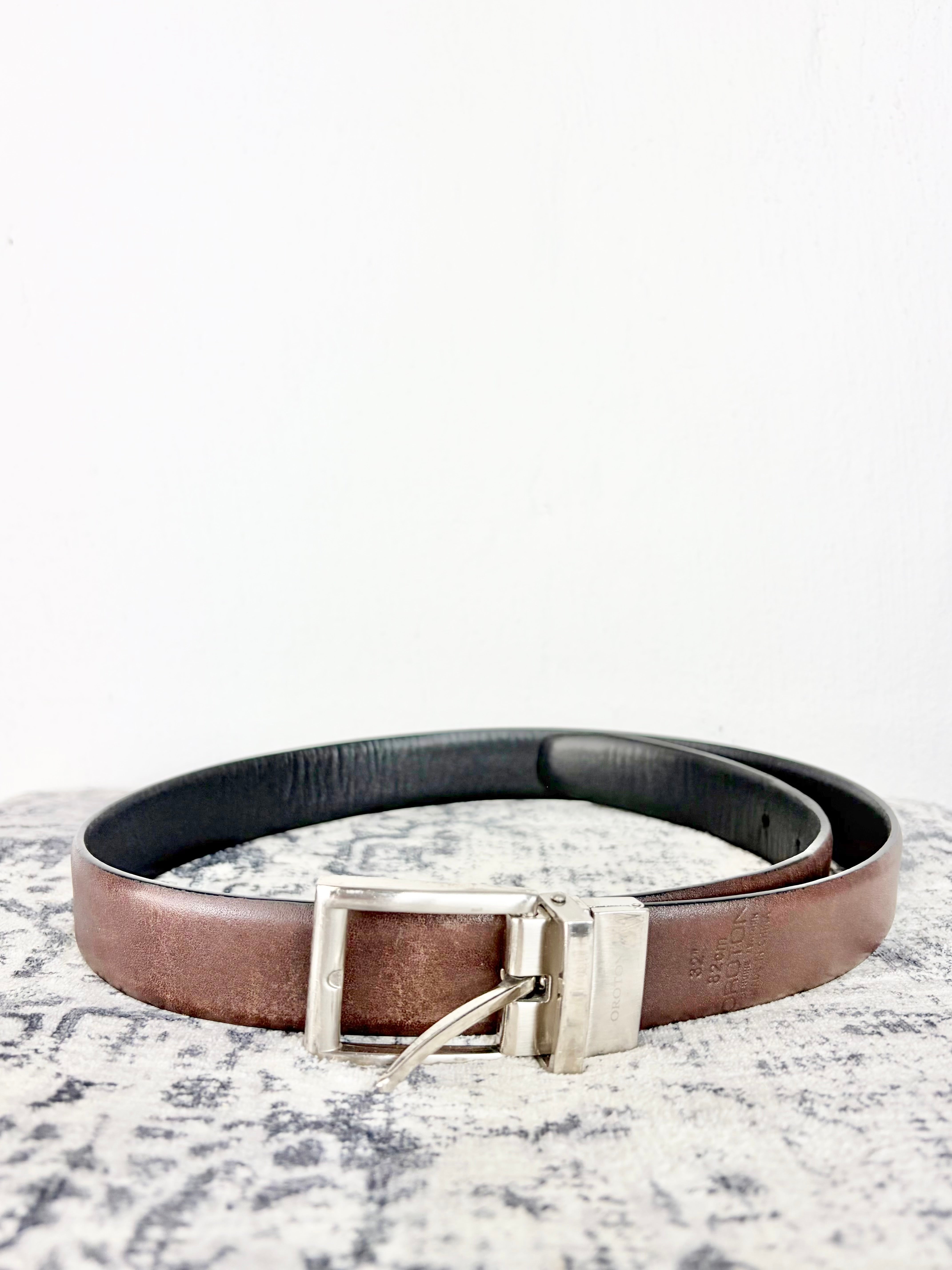 A brown leather belt with a silver-toned metal buckle, coiled on a textured gray and white surface against a plain white background.