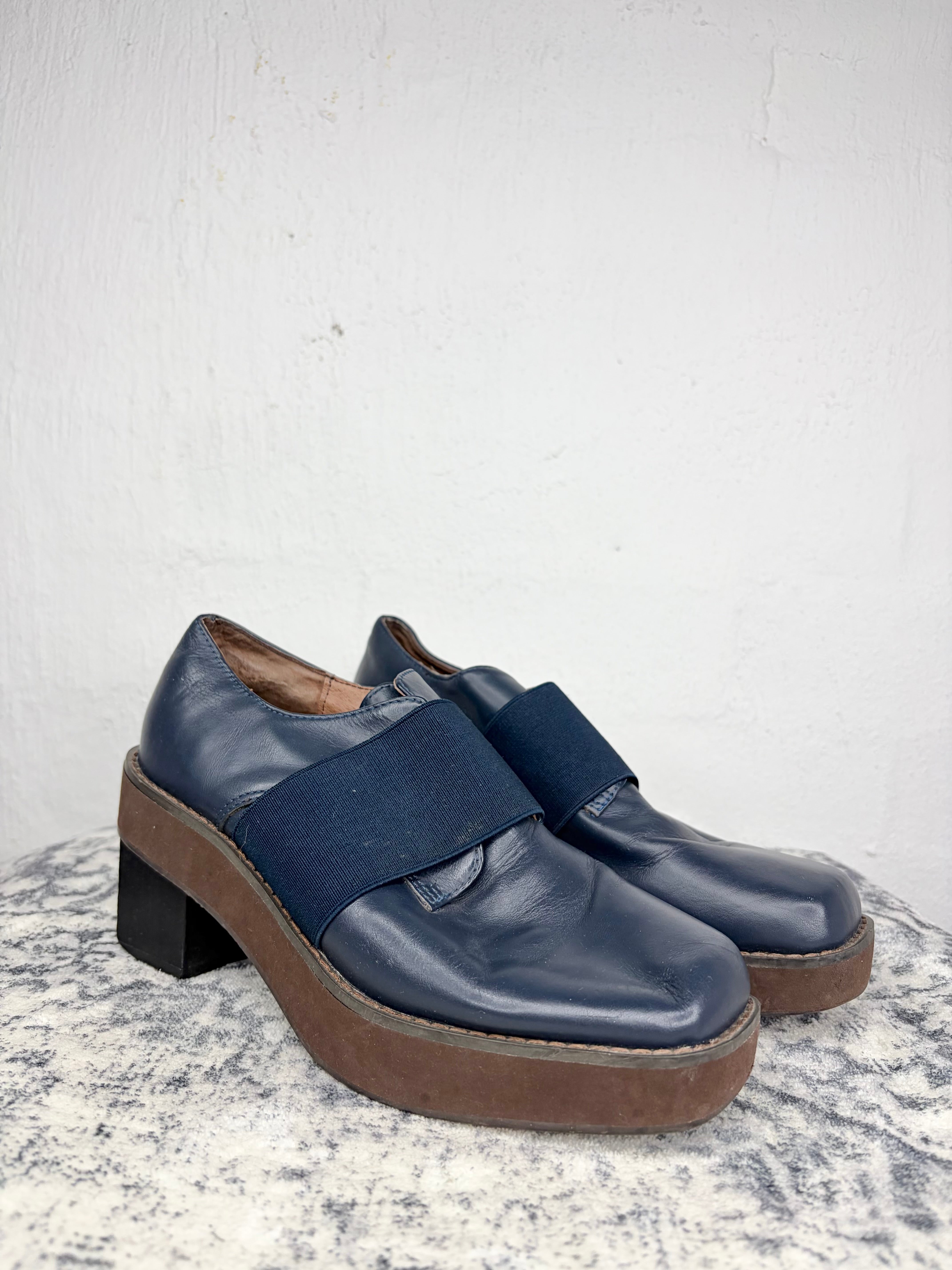 A pair of navy blue leather platform shoes with wide elastic straps and chunky brown soles, displayed on a textured gray stone surface against a white plaster wall.