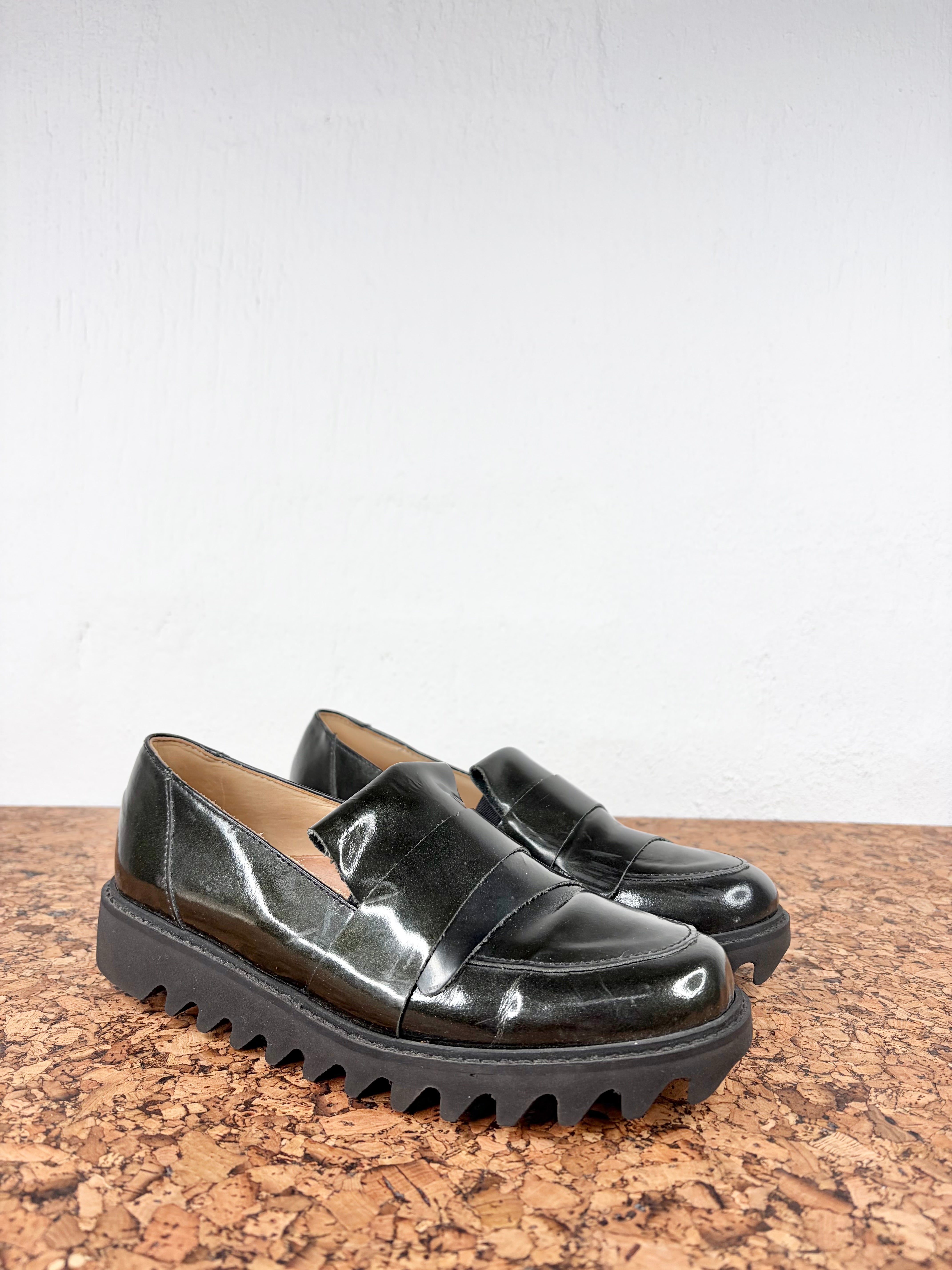 A pair of glossy black patent leather slip-on loafers with chunky lug soles, displayed on a cork surface against a white wall.