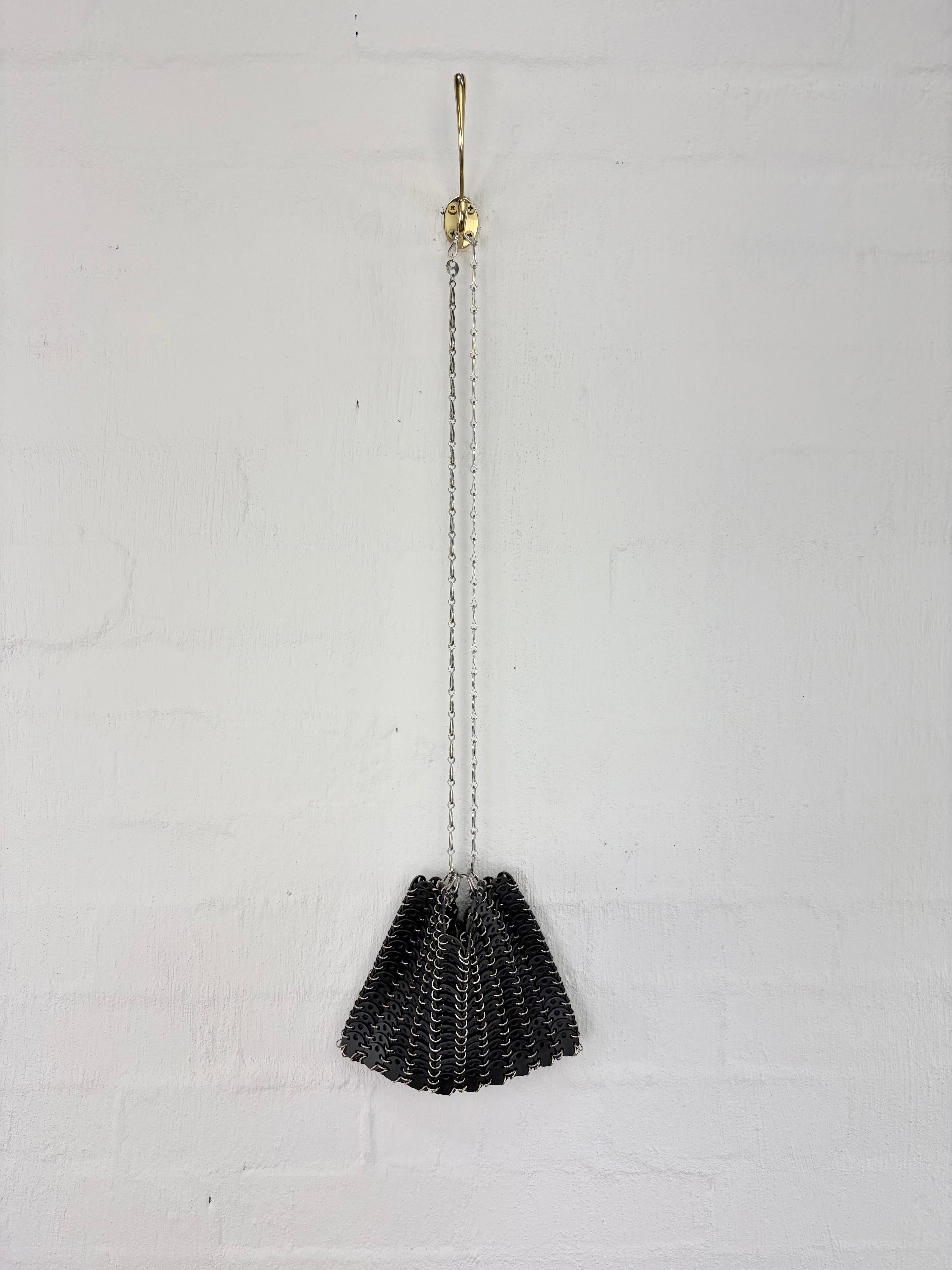 A black woven mesh purse hanging from a gold-toned wall hook with a silver chain, set against a white painted brick wall.