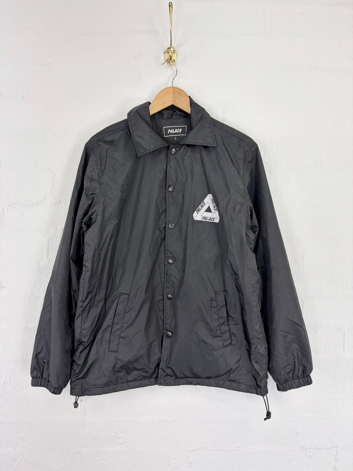 A black Palace Coach jacket hanging on a wooden hanger against a white brick wall. The jacket features the iconic Palace Tri-Ferg logo on the left chest and a black label on the collar.