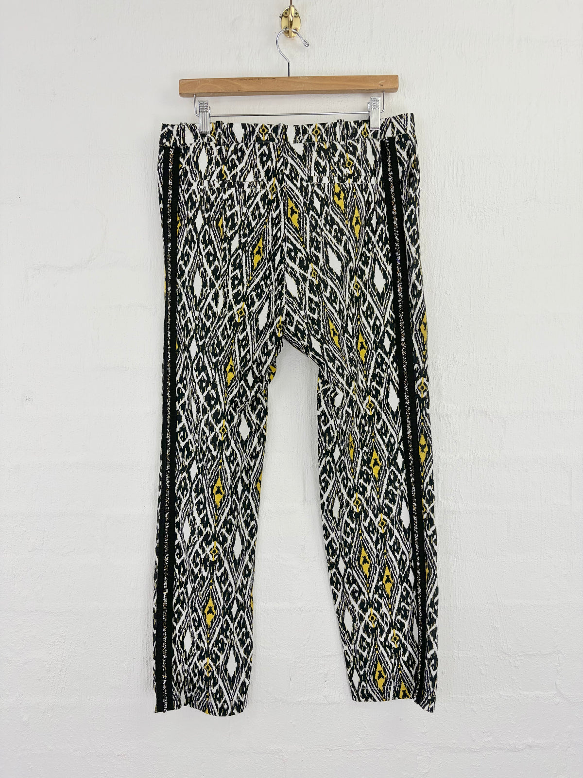 Black, white, and yellow geometric patterned pants hanging from a wooden hanger against a white brick wall.