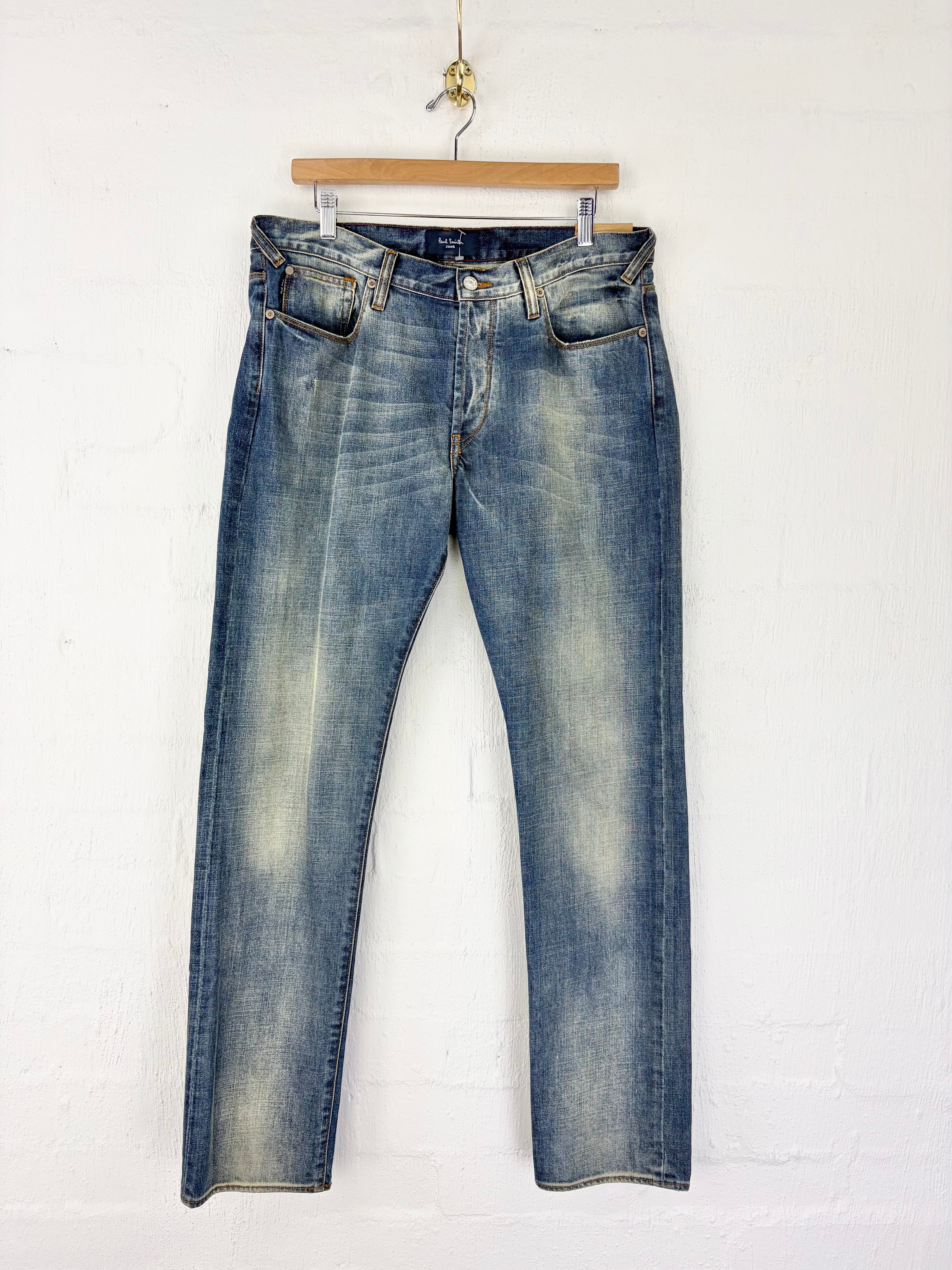 A pair of blue denim jeans with a faded wash, hanging from a wooden hanger against a white painted brick wall.