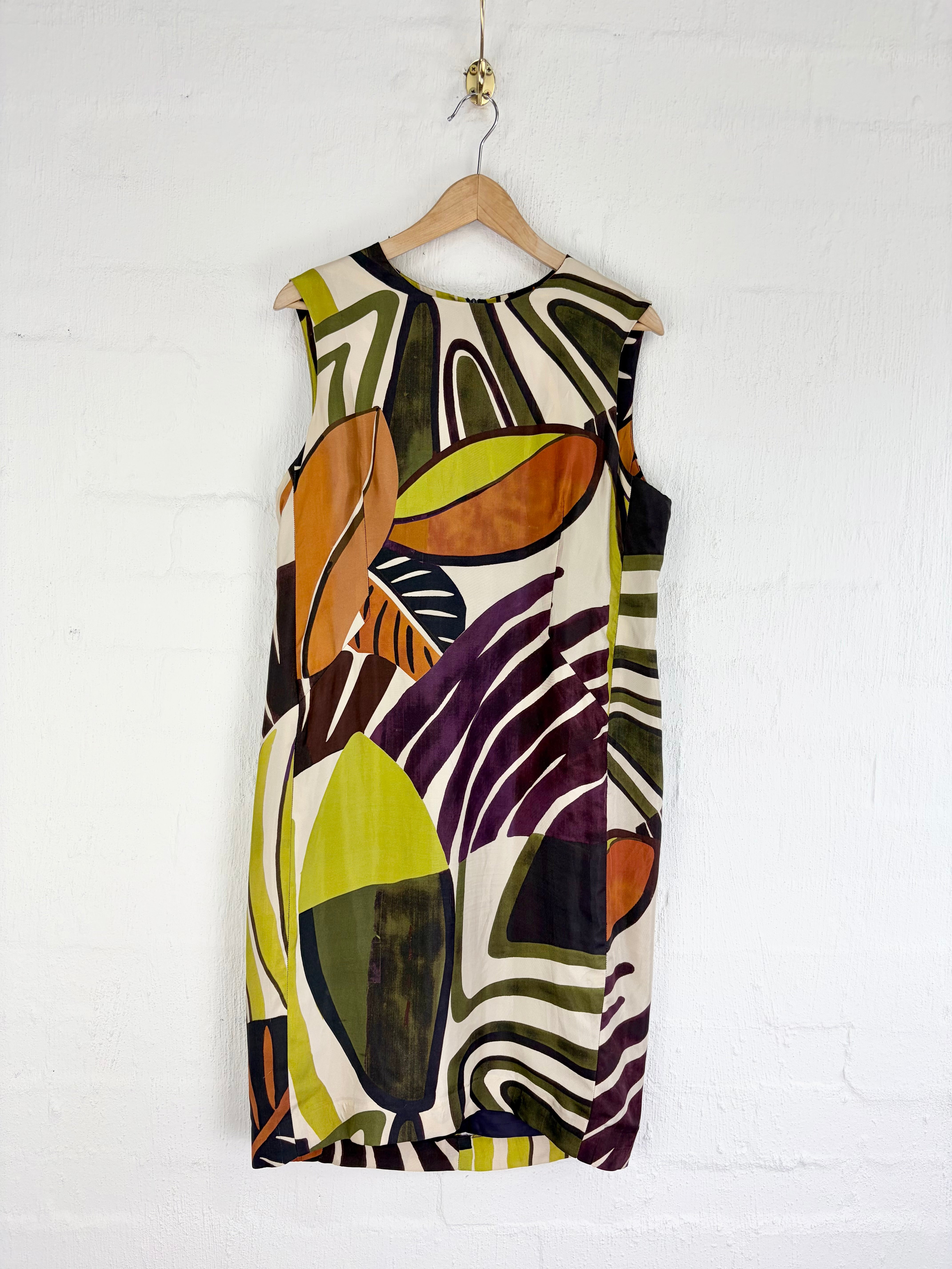 A sleeveless knee-length dress with an abstract geometric print in olive green, mustard yellow, orange, purple, black, and cream, hanging on a wooden hanger against a white painted brick wall.
