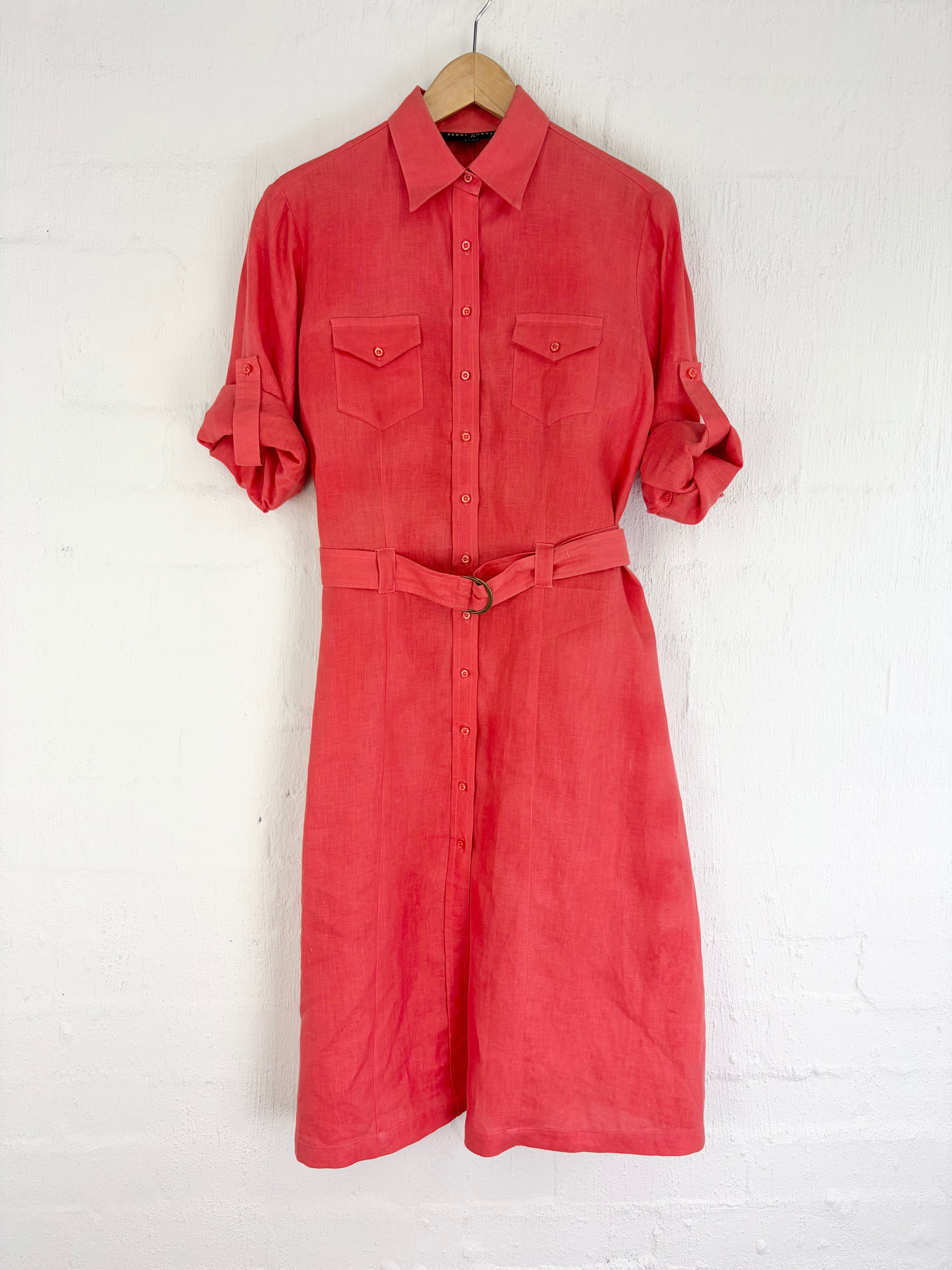 A coral red linen shirt dress with a matching waist belt, hanging on a wooden hanger against a white painted brick wall.