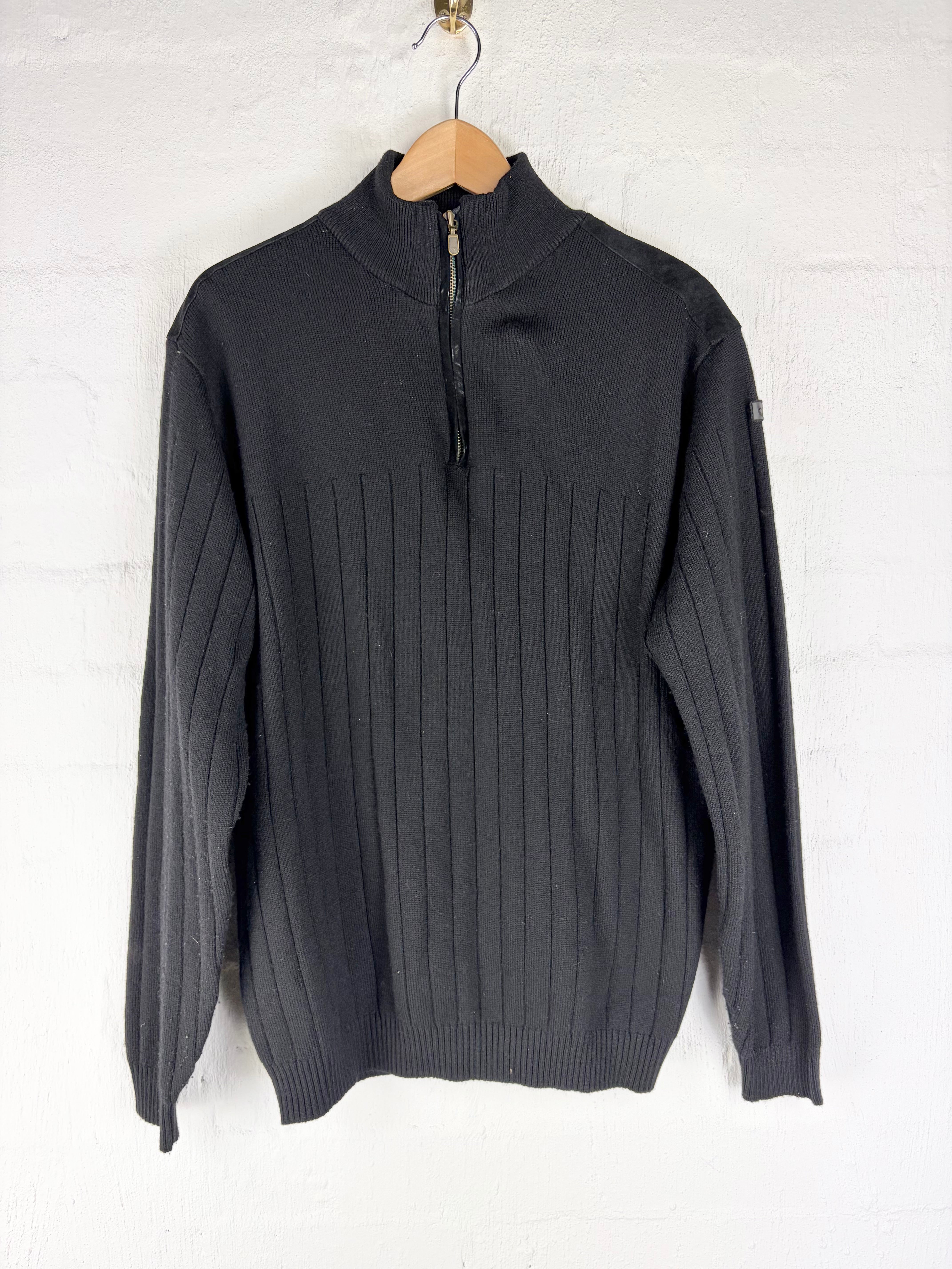 A dark gray ribbed knit quarter-zip sweater hanging on a wooden hanger against a white painted brick wall.
