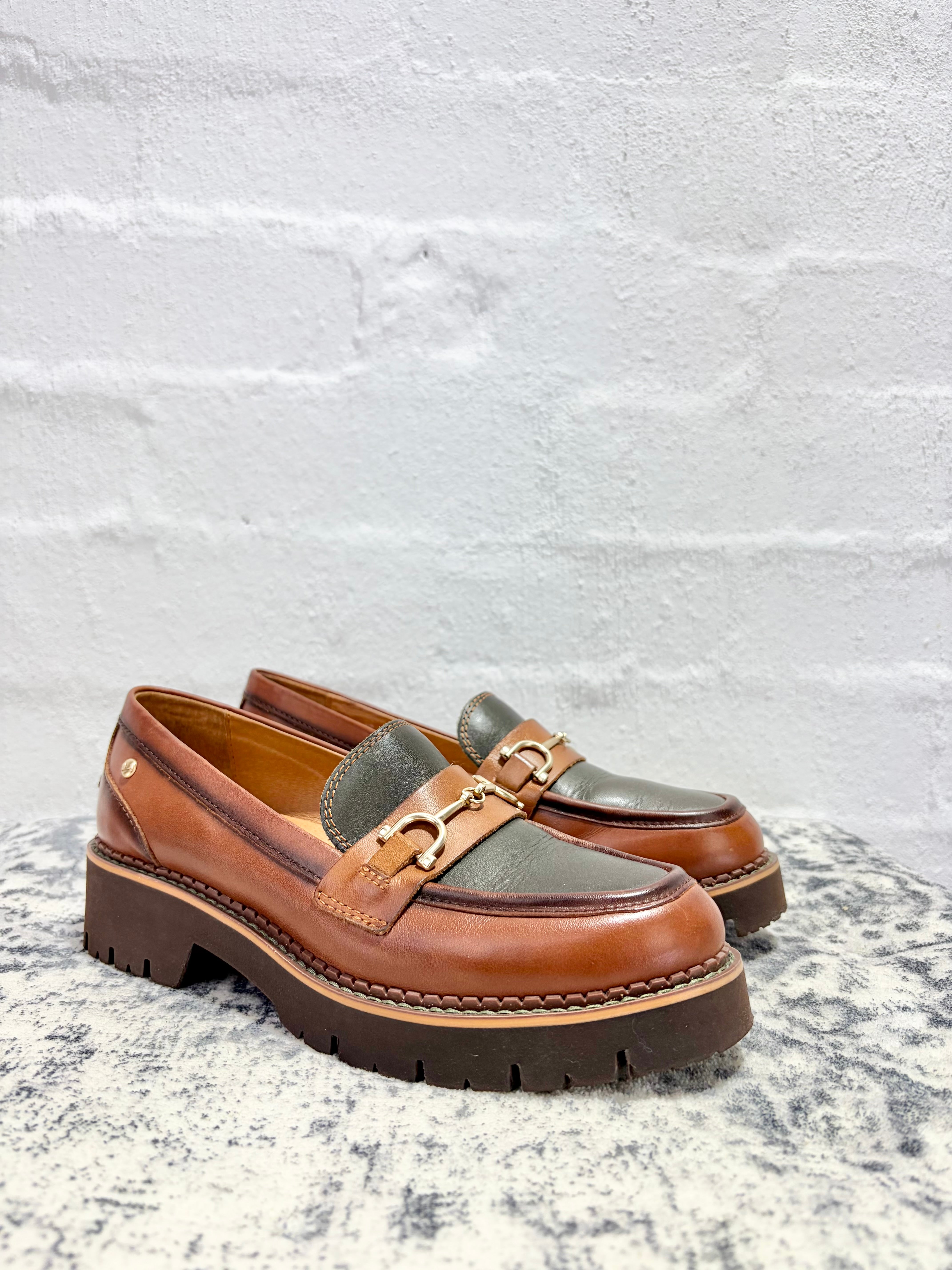 A pair of brown leather platform loafers with dark green suede side panels and a gold-tone horsebit detail, resting on a speckled stone surface against a white brick wall.