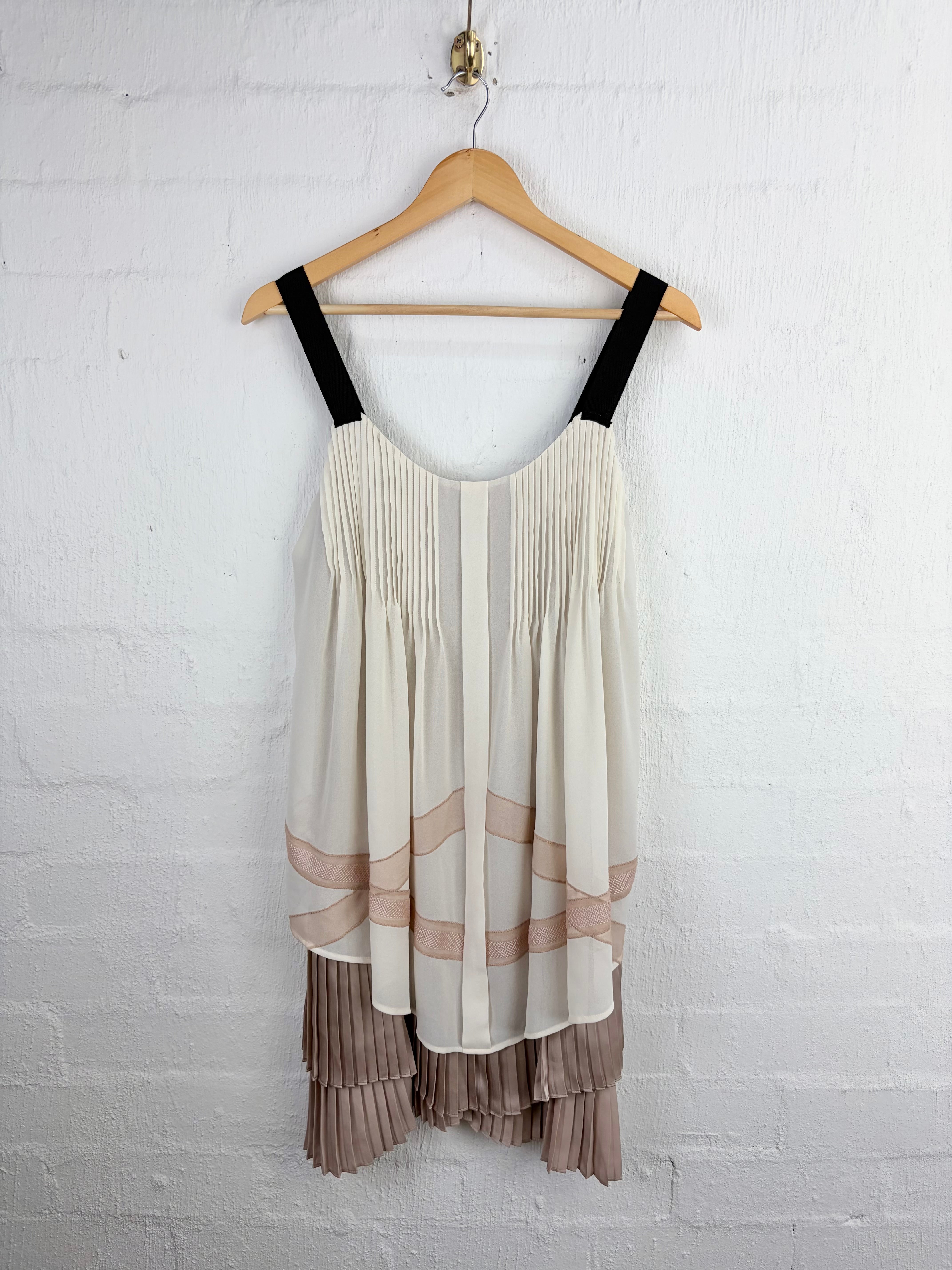 A sleeveless pleated top hanging on a wooden hanger against a white painted brick wall. The top is off-white/cream with black shoulder straps, soft blush-beige horizontal bands through the lower half, and a taupe pleated ruffled hem.