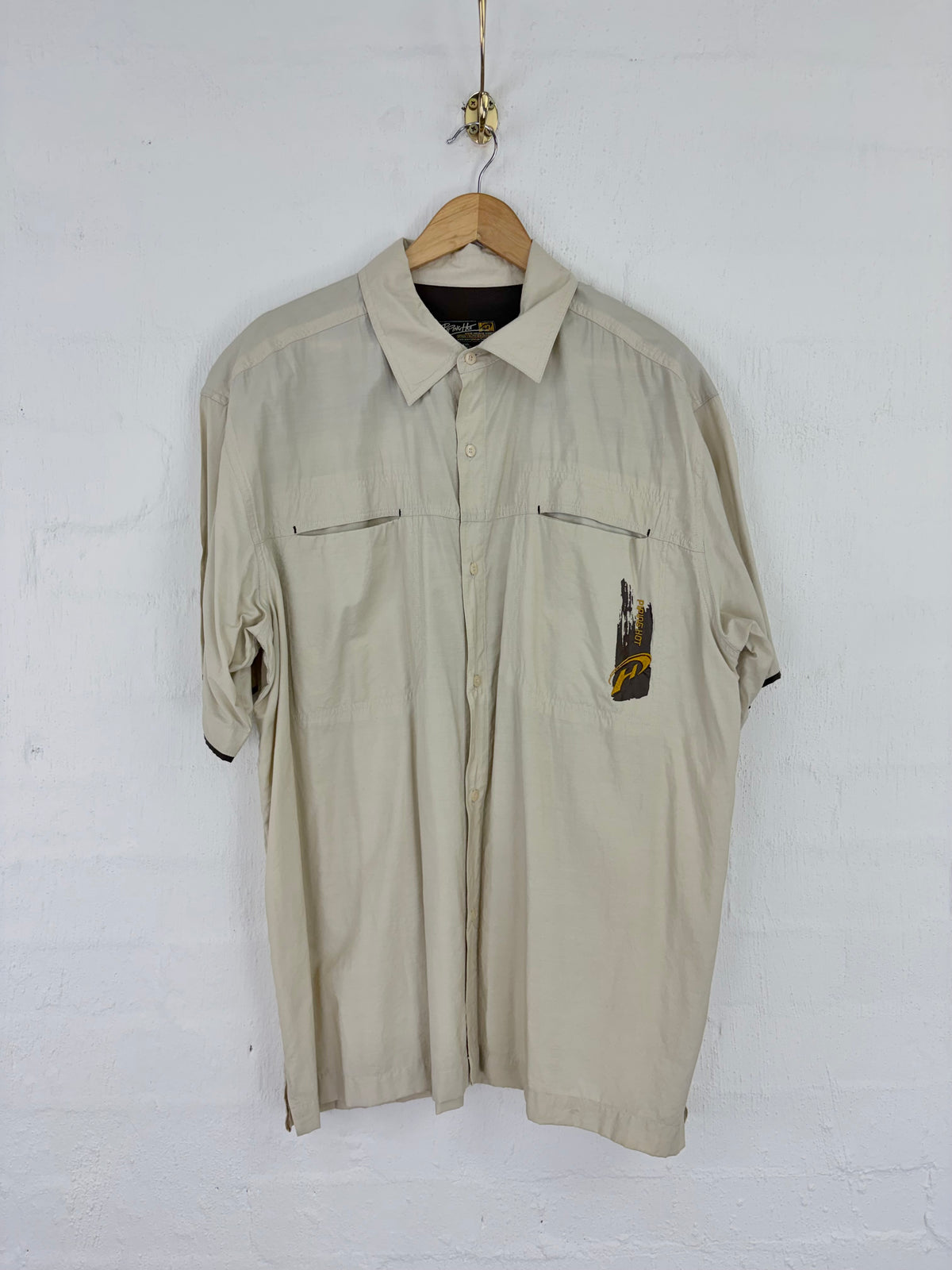 A beige short-sleeve button-up shirt with two chest pockets, hanging on a wooden hanger against a white brick wall. A small black and yellow graphic patch is visible on the left chest pocket.