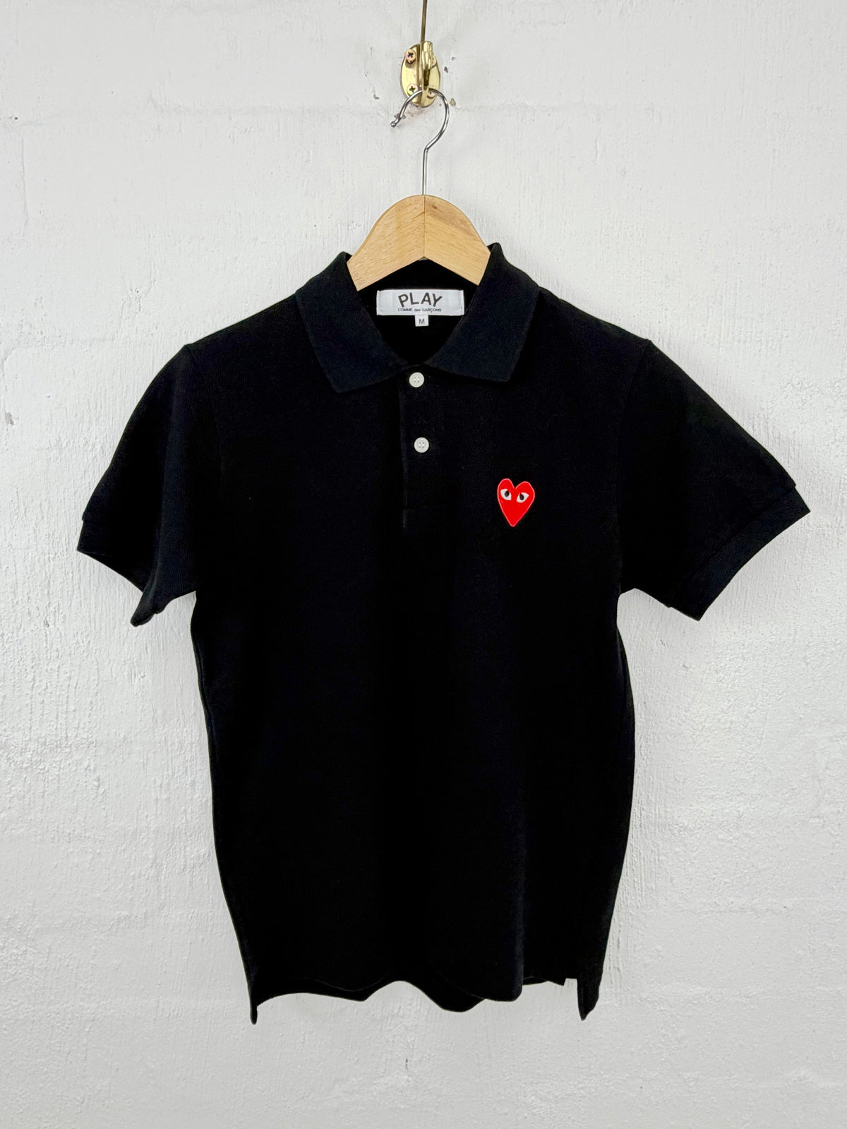 A black short-sleeve polo shirt with a red heart logo on the chest, hanging on a wooden hanger against a white textured wall.