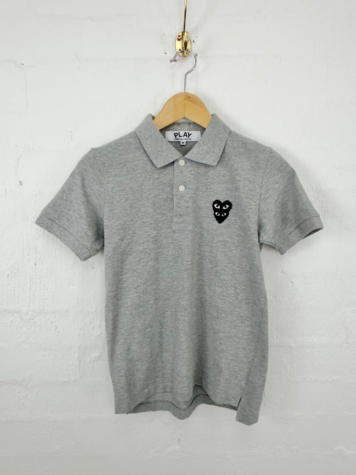 A heather gray short-sleeve polo shirt with a black heart-with-eyes logo on the left chest, hanging on a wooden hanger against a white painted brick wall.