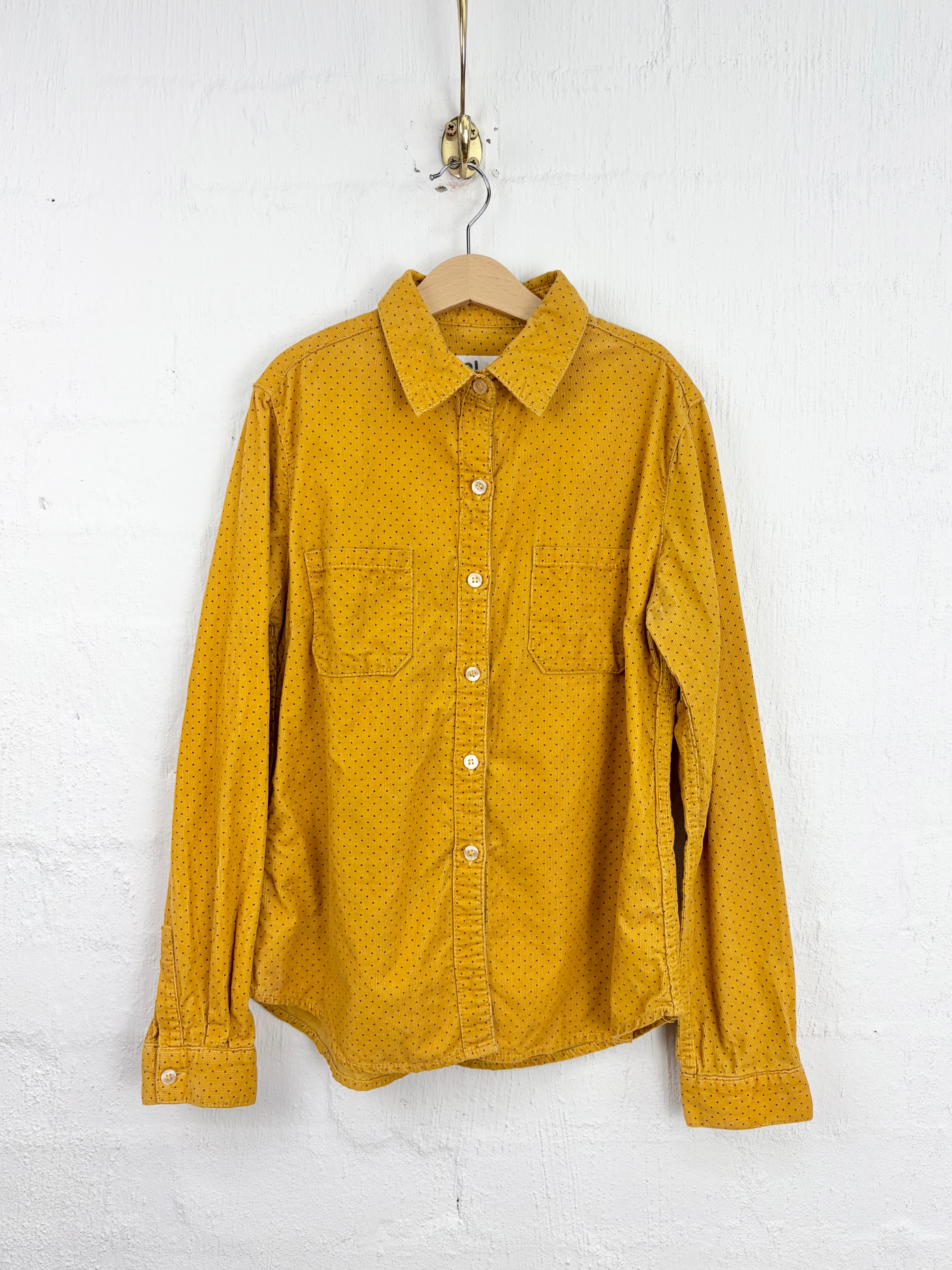 A mustard yellow long-sleeve button-up shirt with a subtle micro-pattern, hanging on a wooden hanger against a white textured wall.