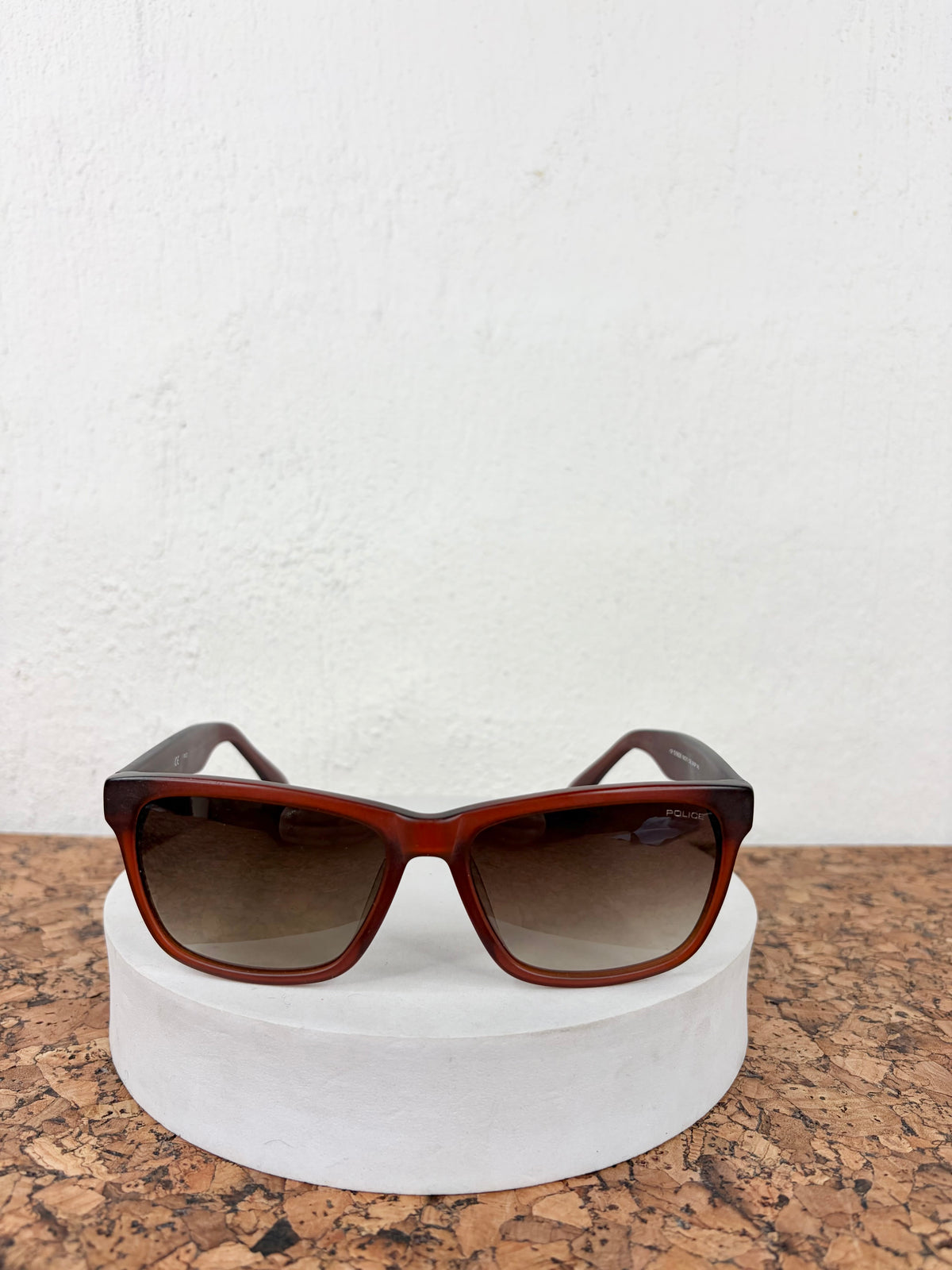 Rustic brown acetate square sunglasses with gradient brown lenses, displayed on a white cylindrical pedestal over a cork surface against a white textured wall.