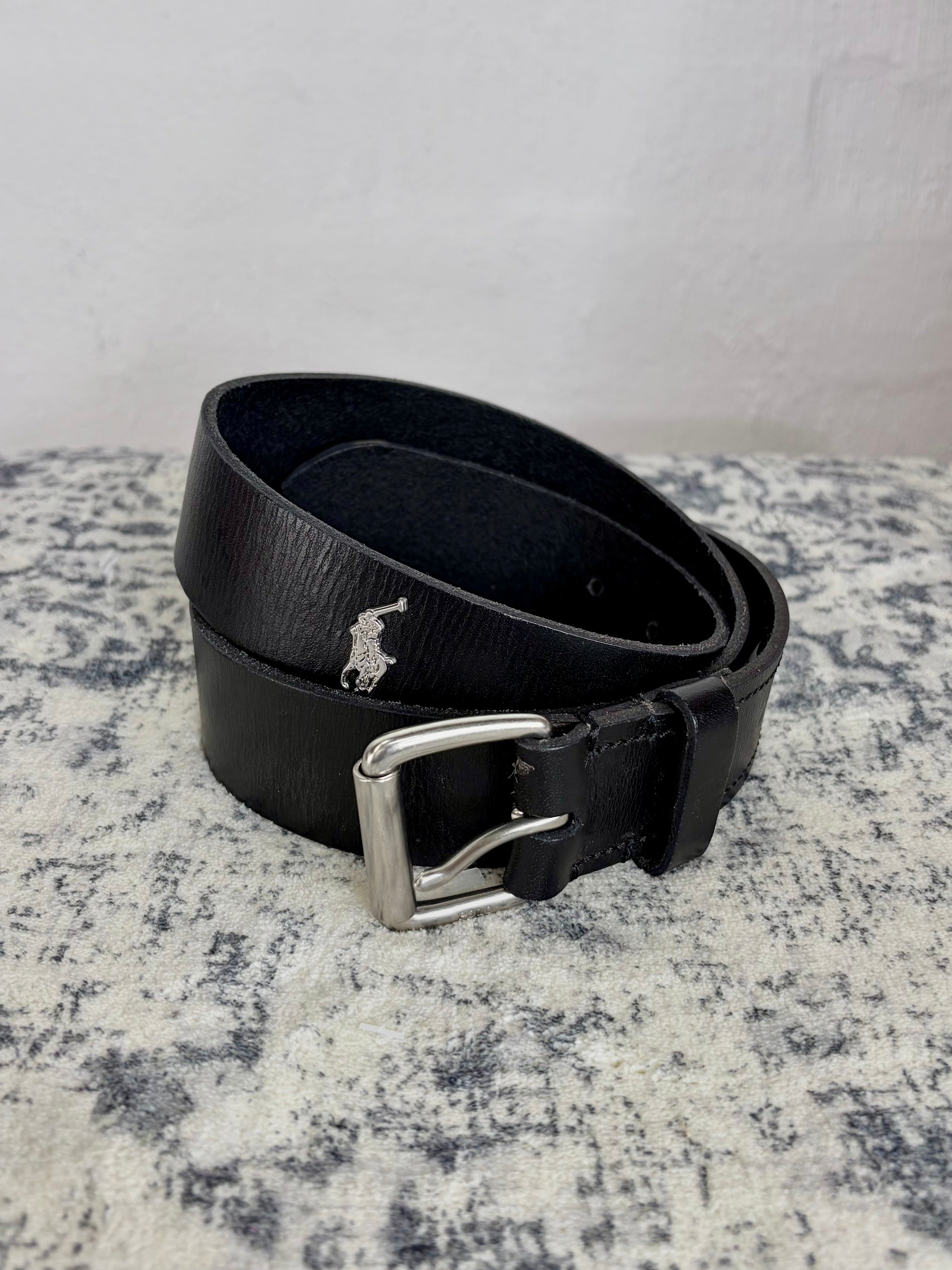 A black leather belt with a silver-tone buckle and a small embroidered polo player logo, coiled on a textured gray and white surface against a light gray wall.