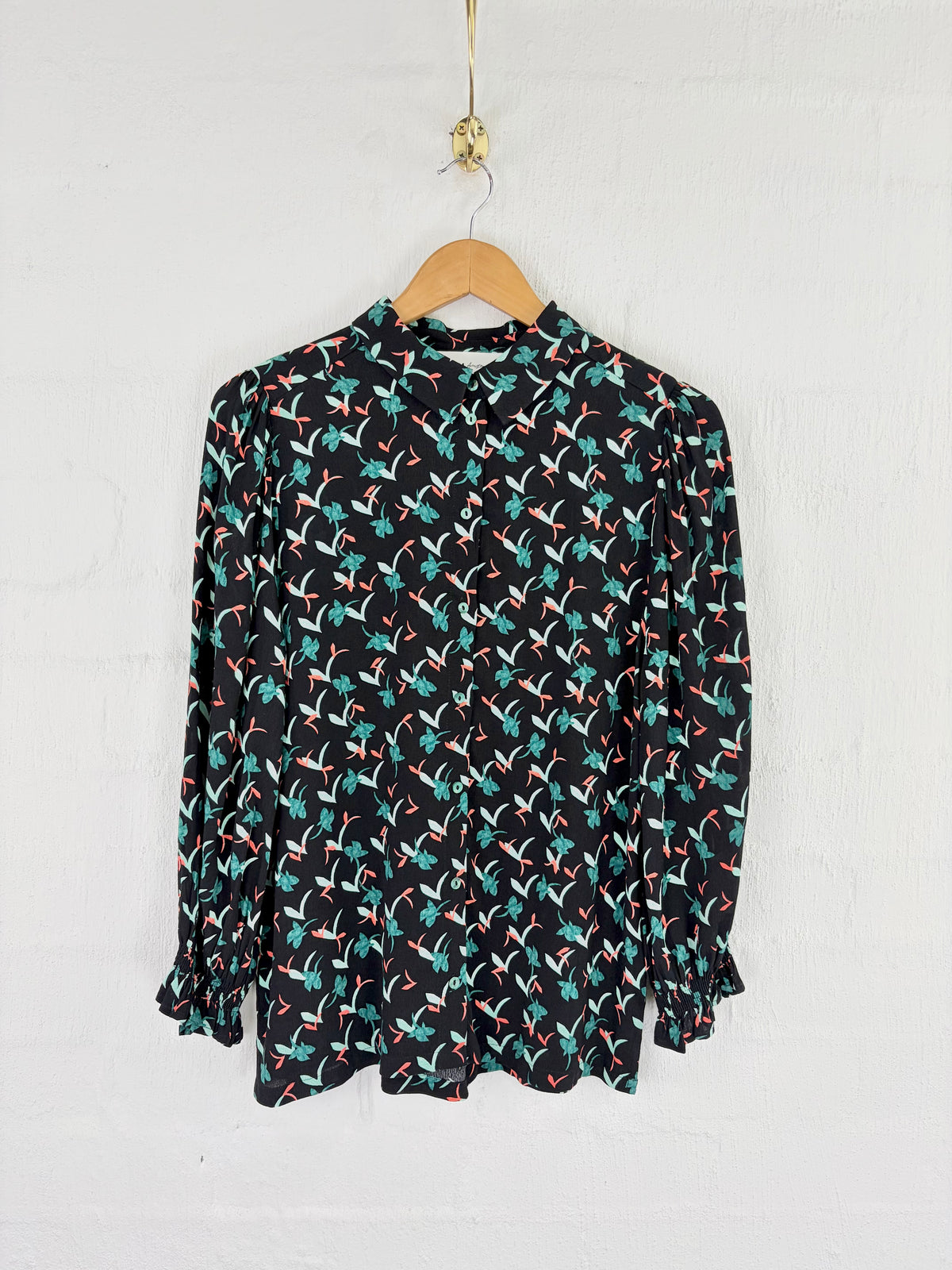 A long-sleeve blouse with a black base and an all-over abstract print in teal, orange, and white, hanging on a wooden hanger against a white brick wall.