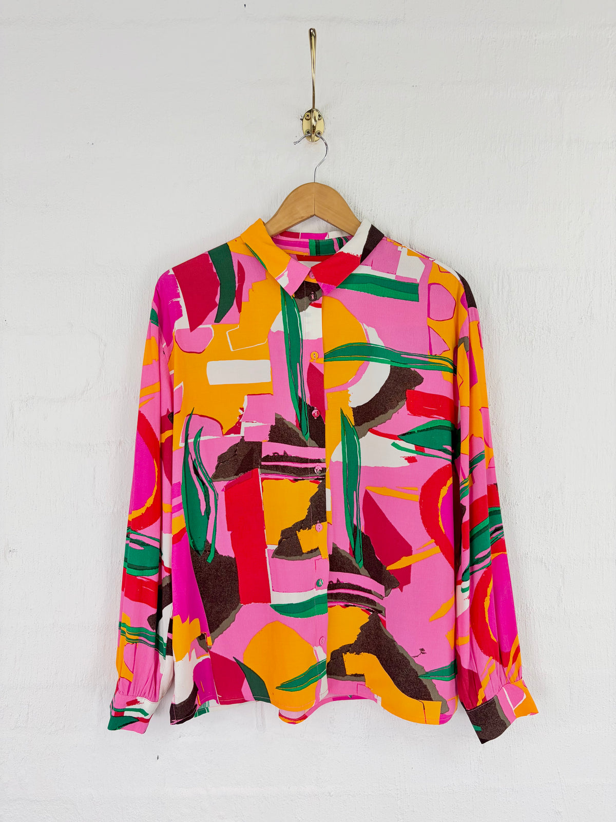 A long-sleeve button-up shirt with a bold abstract geometric print in pink, orange, yellow, green, and brown, hanging on a wooden hanger against a white painted brick wall.