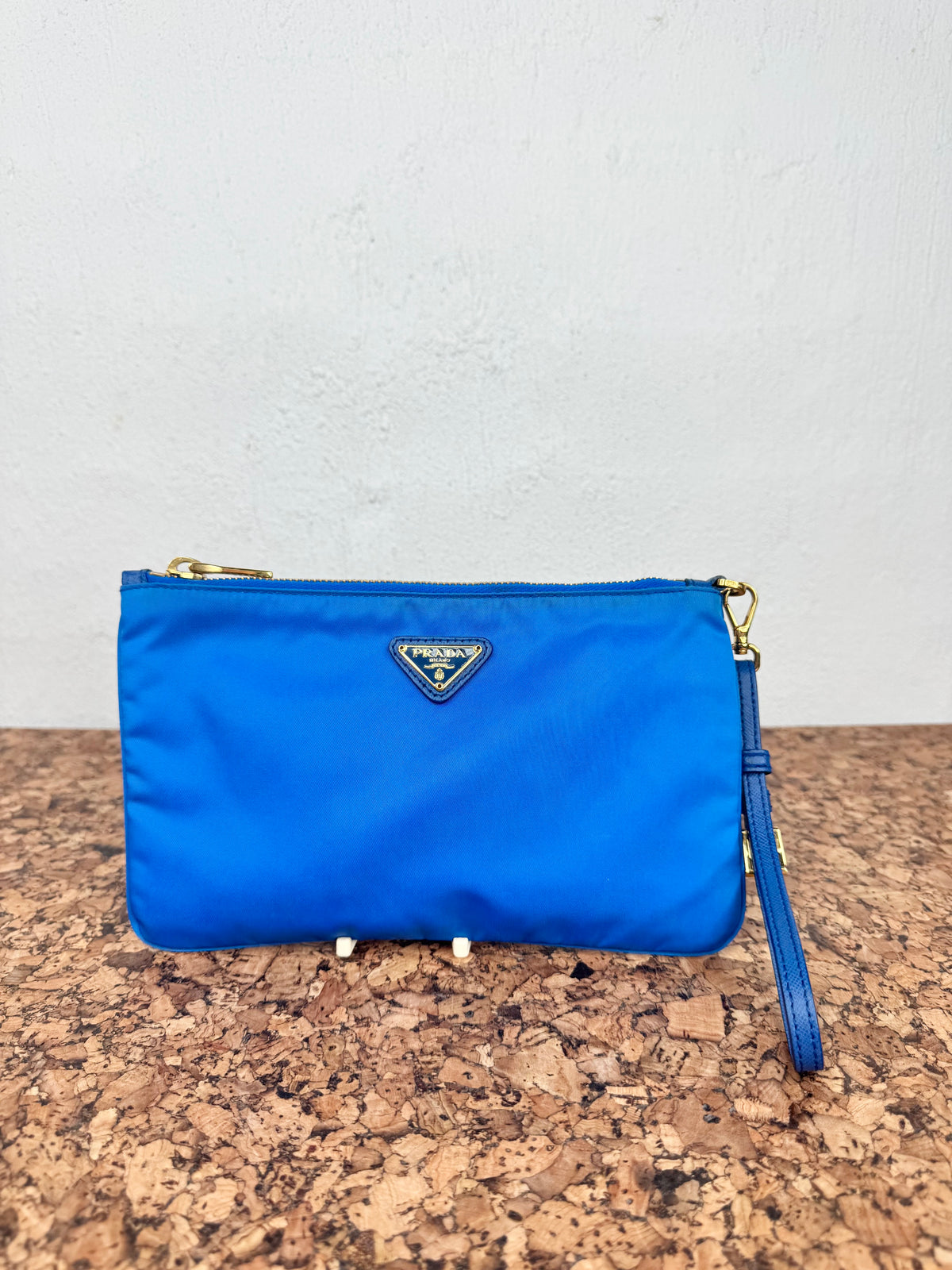 A blue Prada nylon wristlet clutch with gold-tone hardware, resting on a cork surface against a white wall.