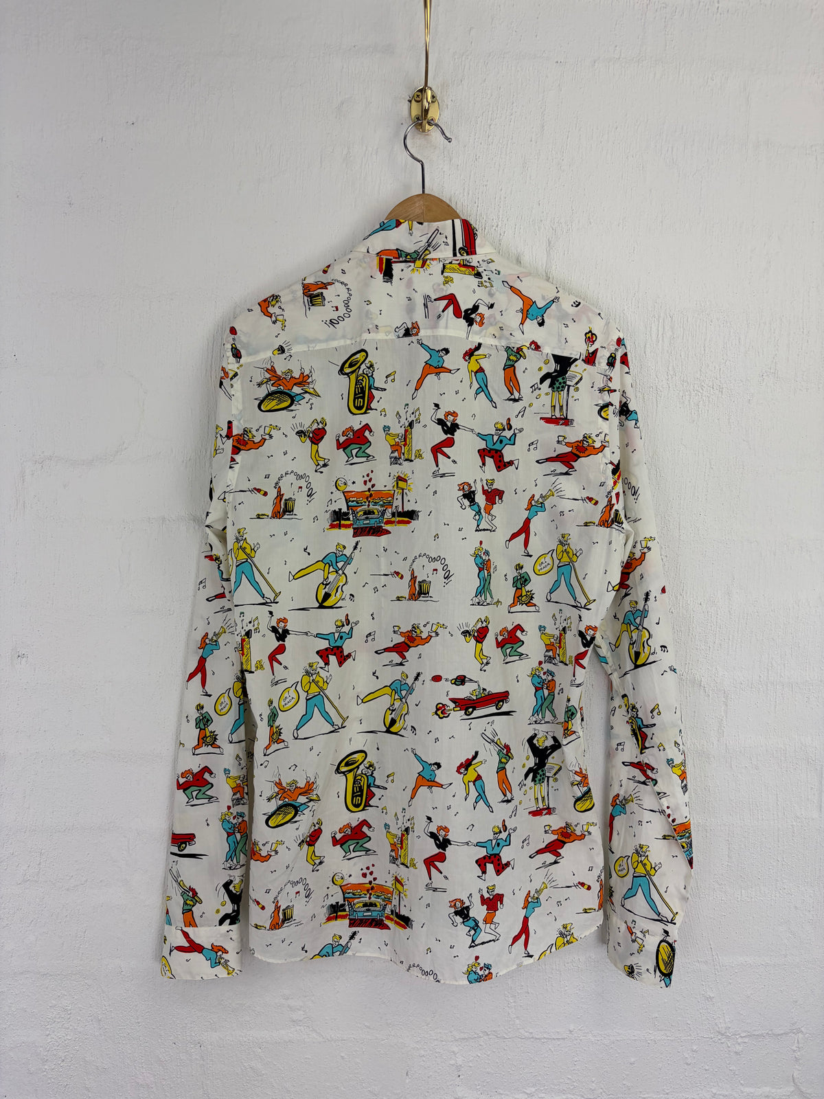 A long-sleeve button-up shirt with a colorful all-over cartoon print of figures, sports, and objects on an off-white background, hanging on a wooden hanger against a white painted brick wall.