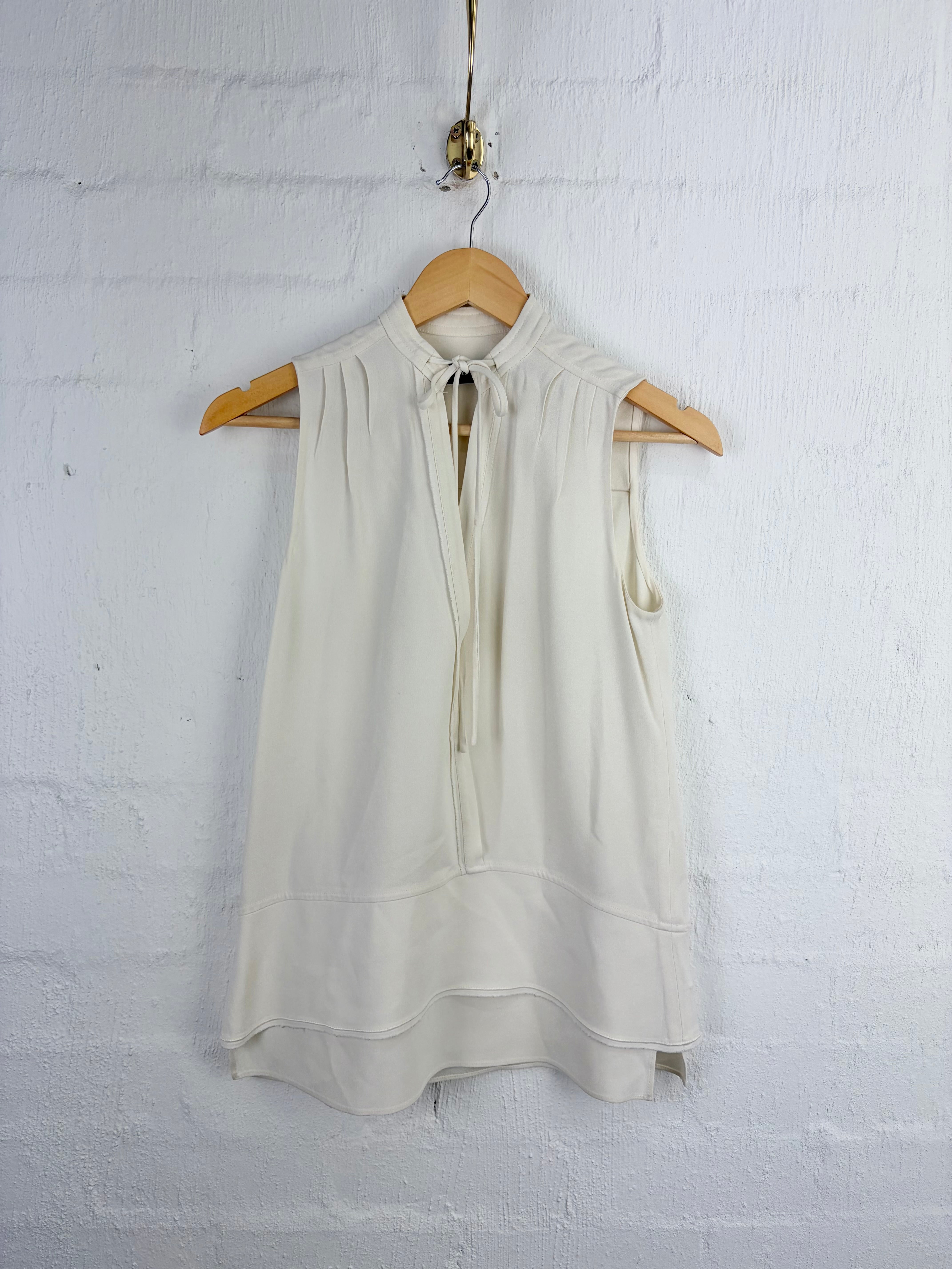 An off-white sleeveless blouse with a tie-neck detail, hanging on a wooden hanger against a white painted brick wall.