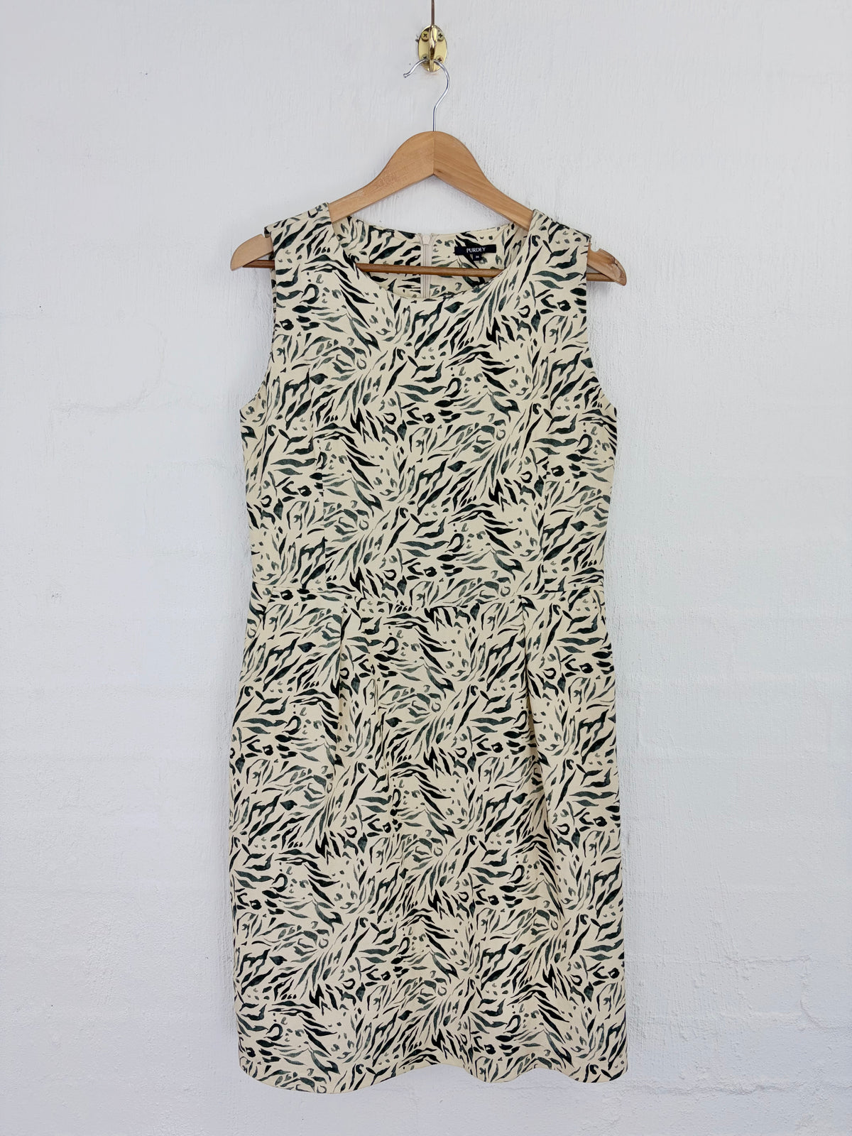 A sleeveless knee-length dress with an abstract black and beige brushstroke pattern, hanging on a wooden hanger against a white painted brick wall.