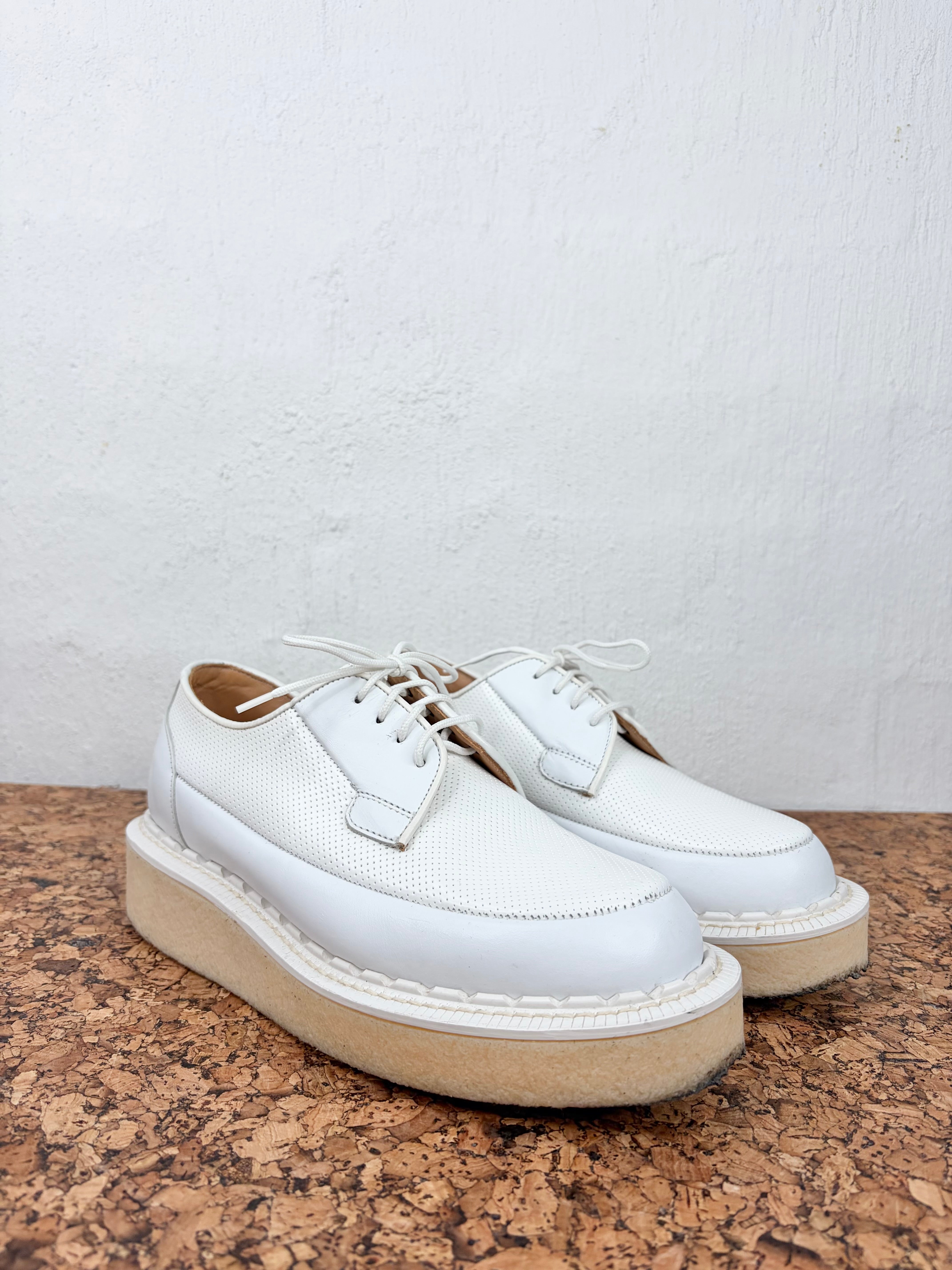 A pair of white leather lace-up shoes with chunky beige platform soles, displayed on a brown cork surface against a white textured wall.