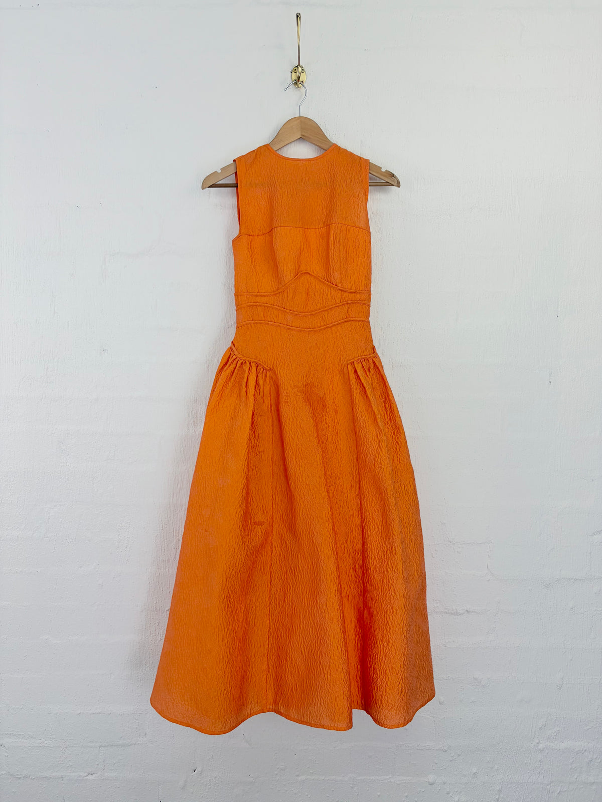 An orange sleeveless midi dress with a textured fabric, hanging on a wooden hanger against a white painted brick wall.