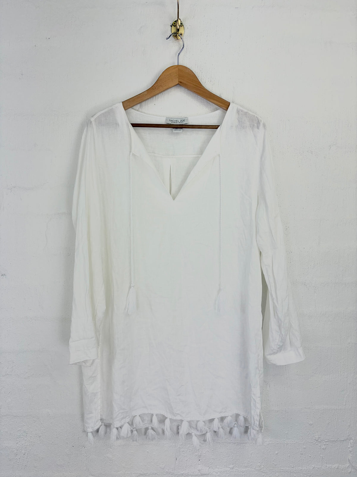 A white long-sleeve tunic top with a V-neckline and drawstring details, hanging on a wooden hanger against a white painted brick wall.