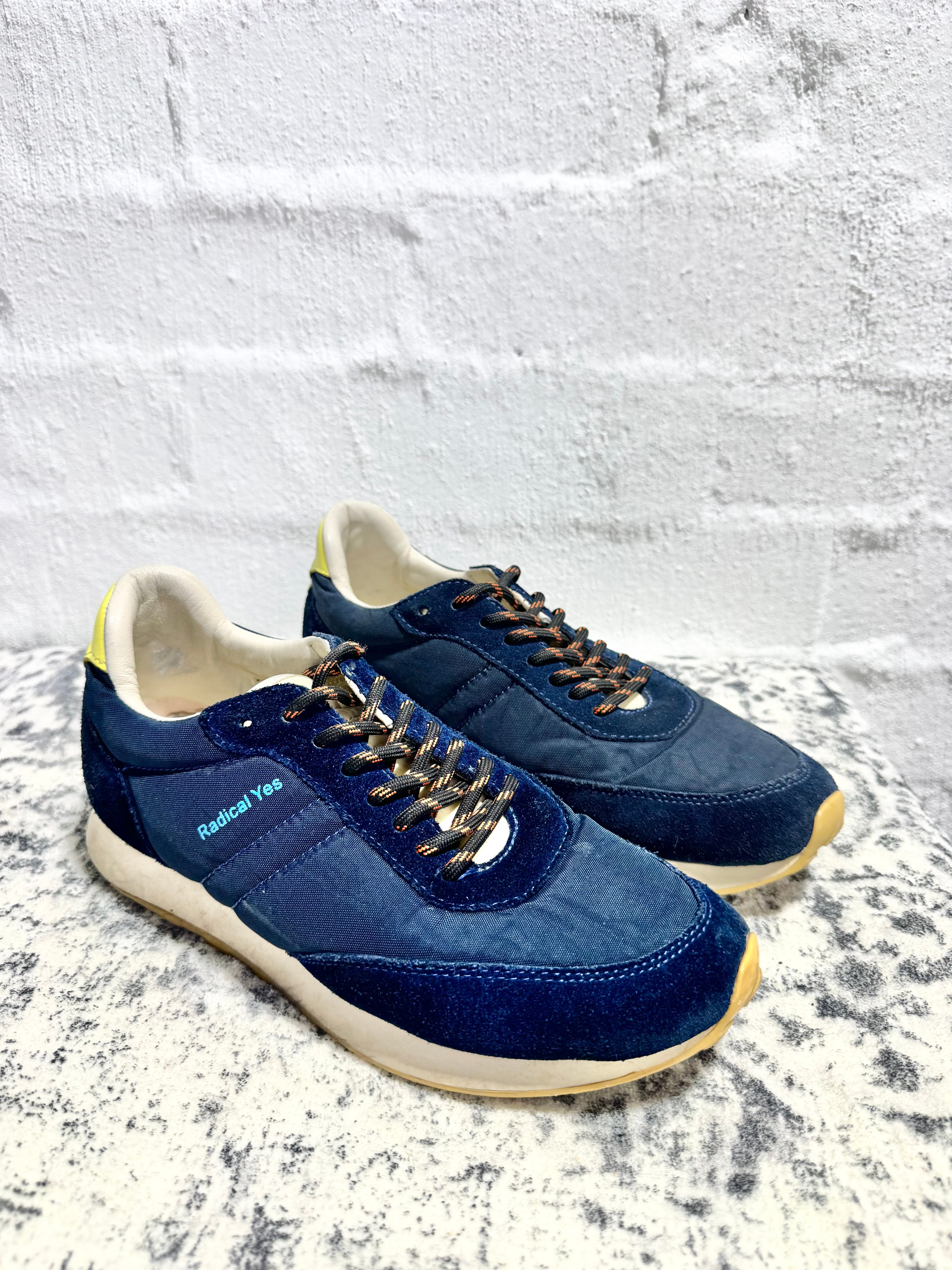 A pair of navy blue suede and canvas sneakers with gold-tone laces and off-white soles, displayed on a textured gray surface against a white brick wall. The brand 'Balenciaga' is visible on the side of the shoe.