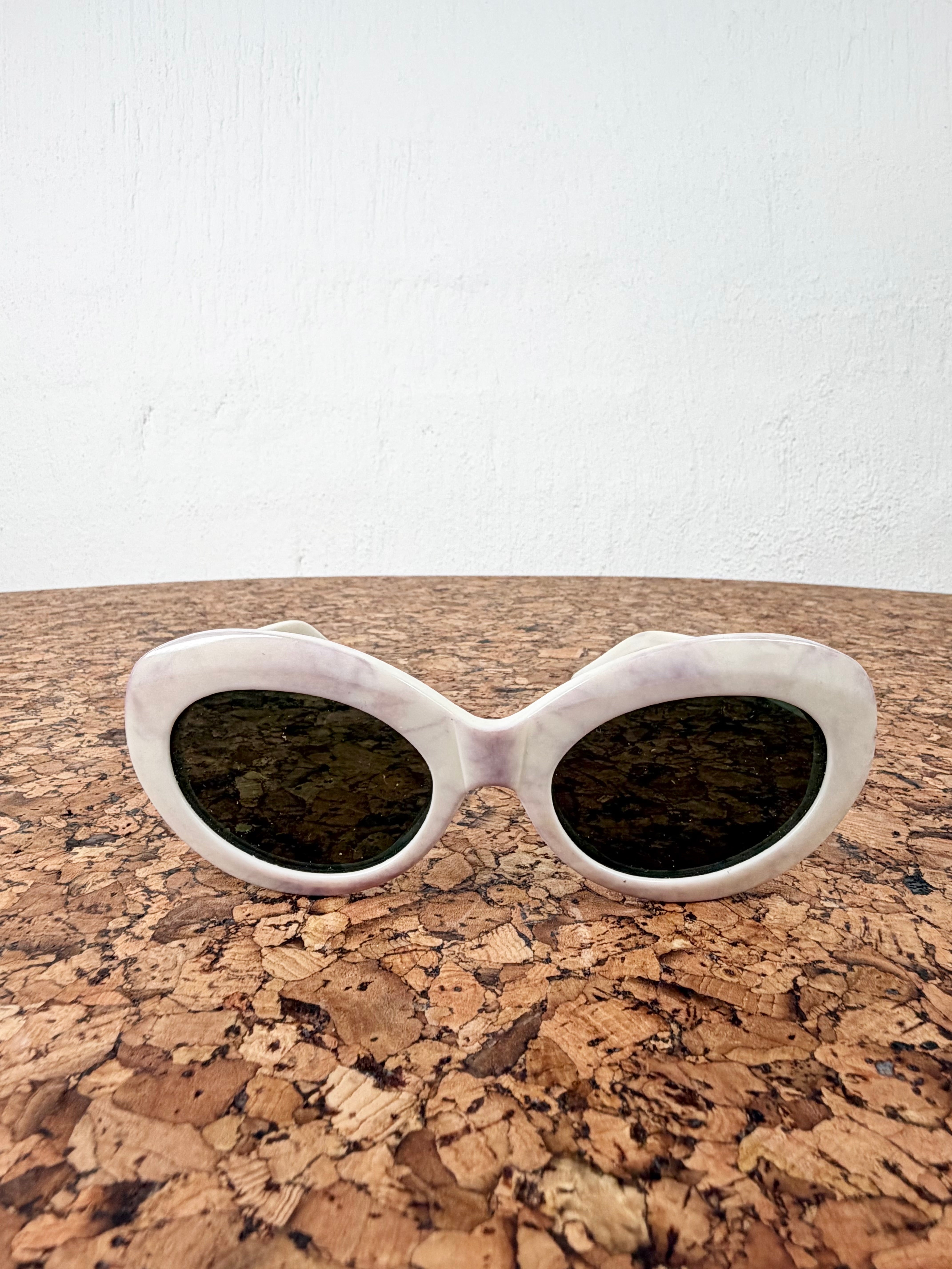 Oval sunglasses with a chunky white frame and dark tinted lenses, resting on a cork surface against a white wall.