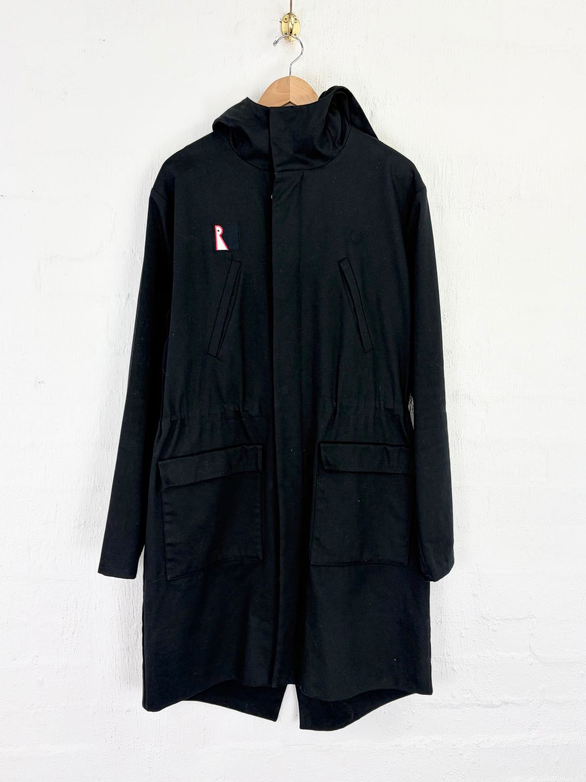 A black hooded jacket with a small pink ribbon graphic on the left chest, hanging on a wooden hanger against a white brick wall.