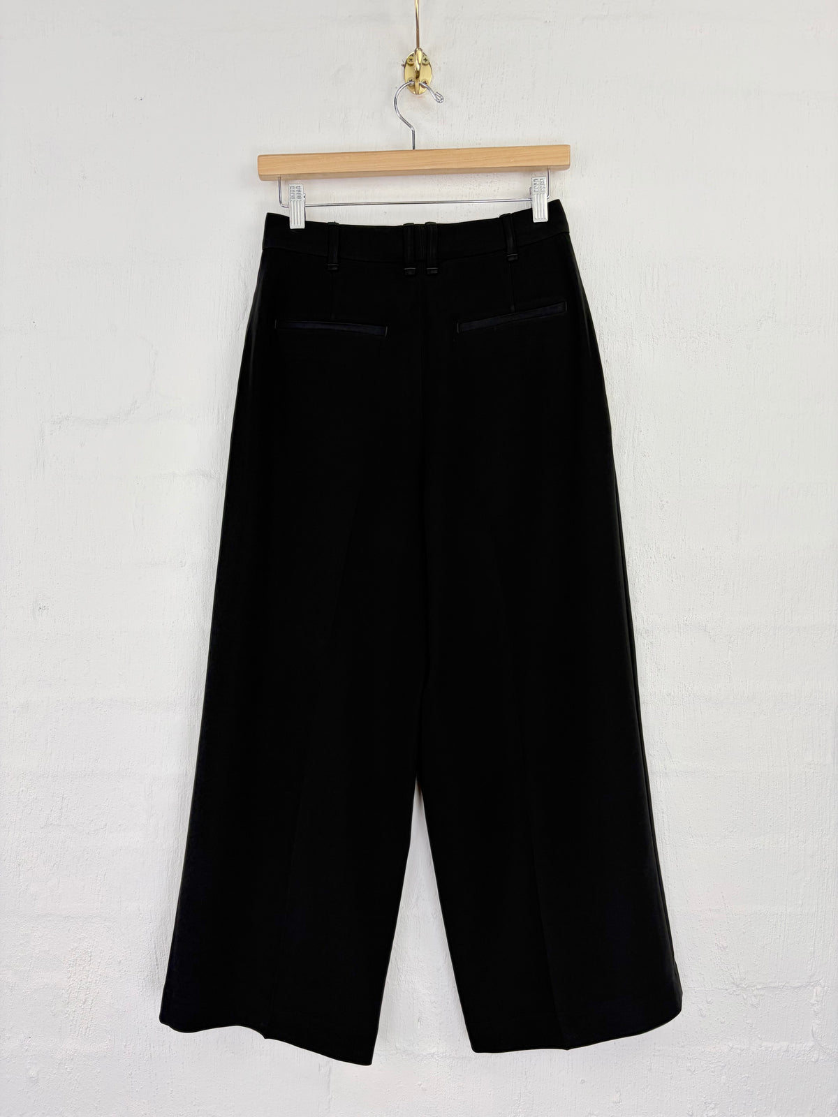 Black wide-leg cropped trousers hanging from a wooden hanger against a white painted brick wall.