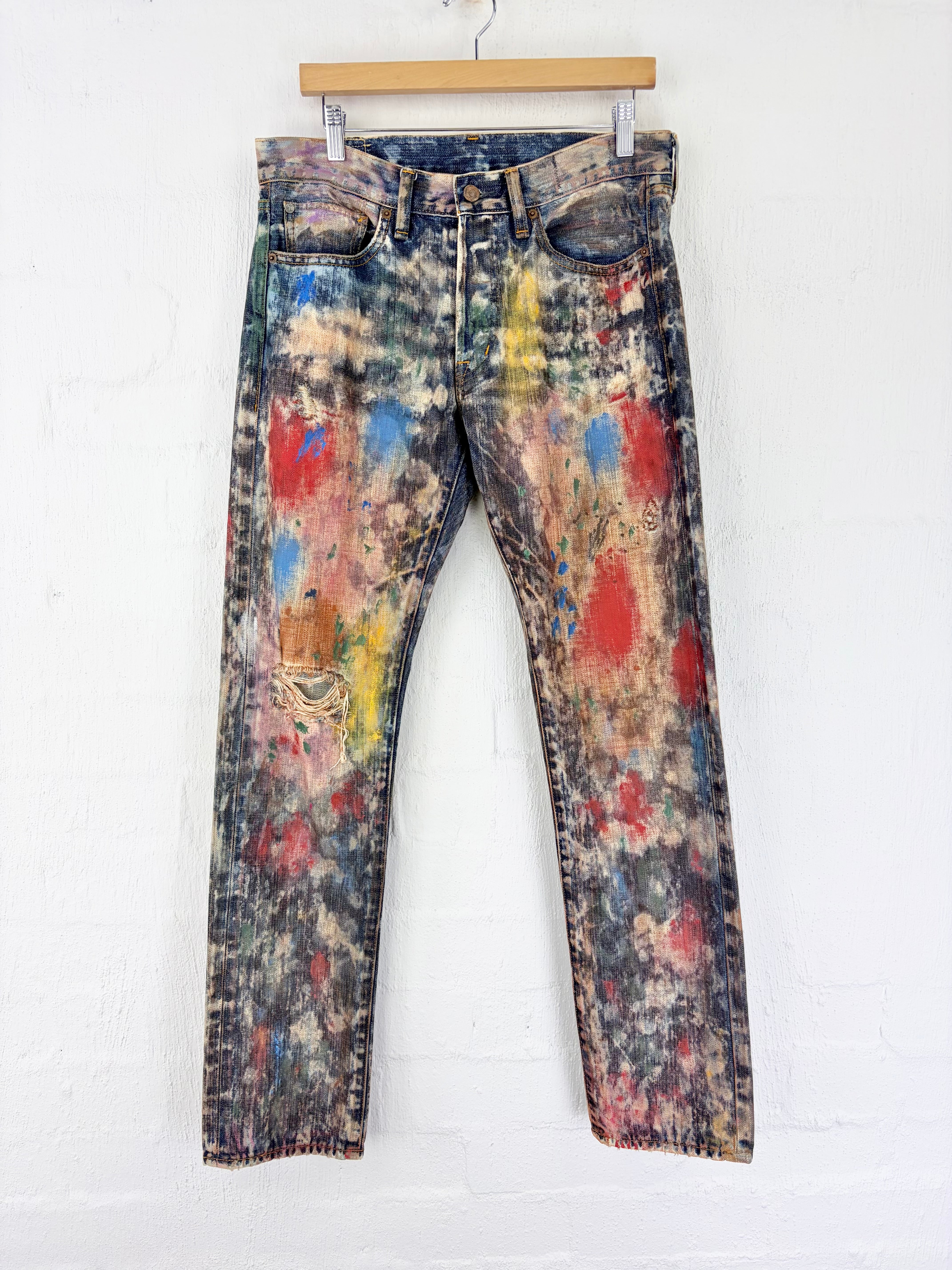 Distressed blue denim jeans with a colorful paint splatter and bleach wash, hanging on a wooden hanger against a white brick wall.