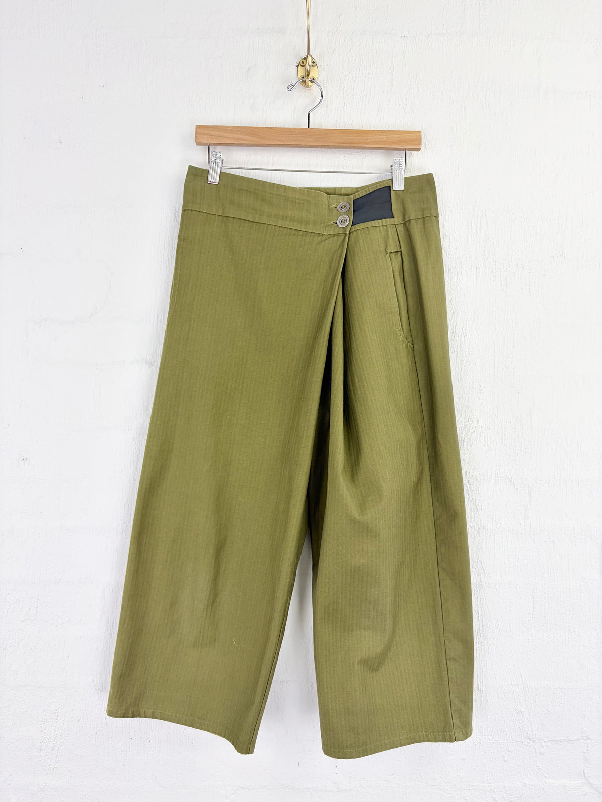 Olive green cropped wide-leg pants hanging on a wooden hanger against a white painted brick wall.