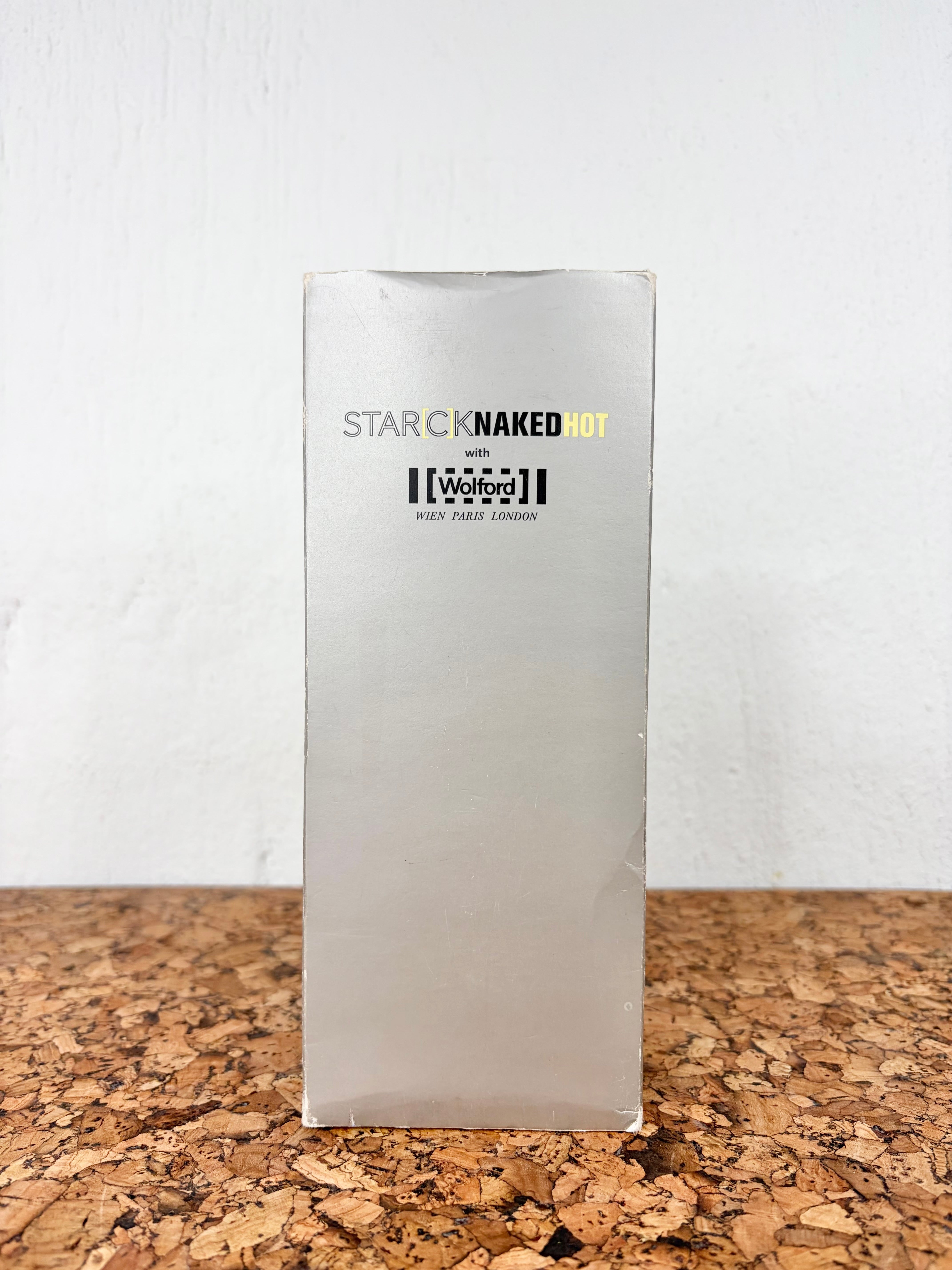 A tall, narrow silver metallic box standing upright on a cork surface against a white wall. The front of the box has the text 'STAR CNAKED HOT' with 'CNAKED' in black and 'HOT' in yellow, plus 'with The Watford' and 'WATford SHIP LONDON' below.