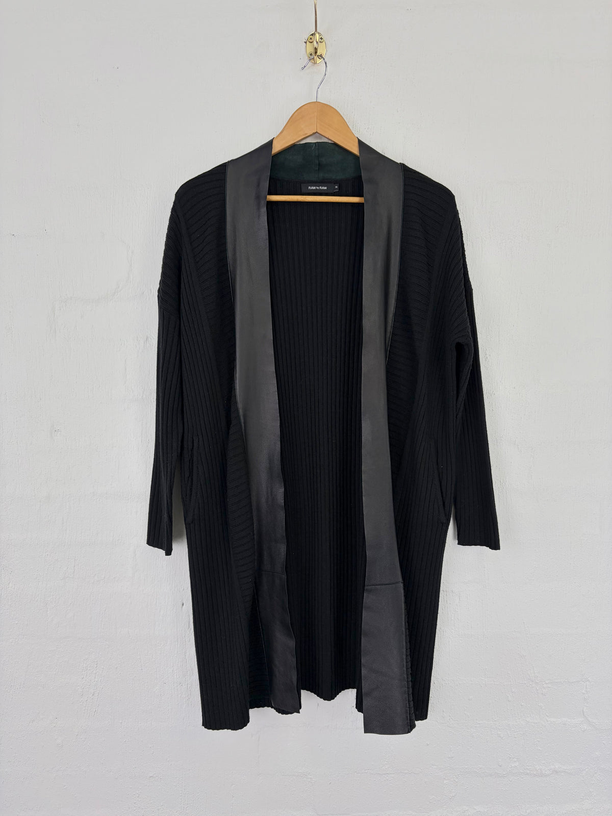 A black open-front cardigan with ribbed texture and wide dark gray satin lapels, hanging on a wooden hanger against a white painted brick wall.
