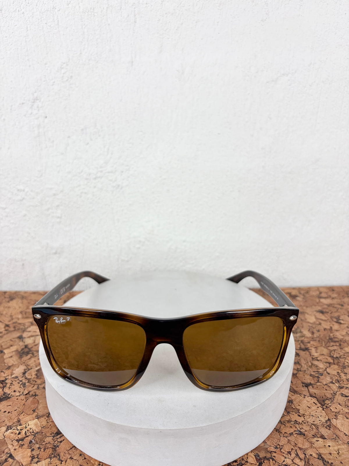 A pair of classic Wayfarer-style sunglasses with a dark brown tortoiseshell frame and amber-tinted lenses, displayed on a white circular pedestal against a white textured wall.
