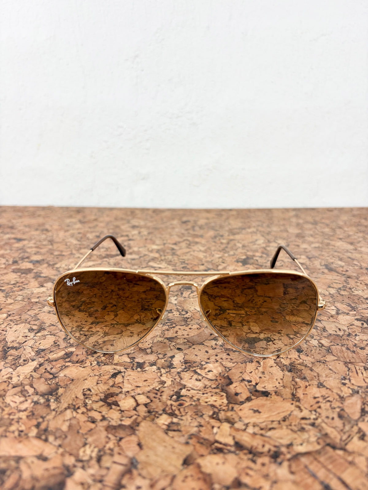 Ray-Ban aviator sunglasses with a gold-tone metal frame and brown gradient lenses, resting on a cork surface against a white wall.