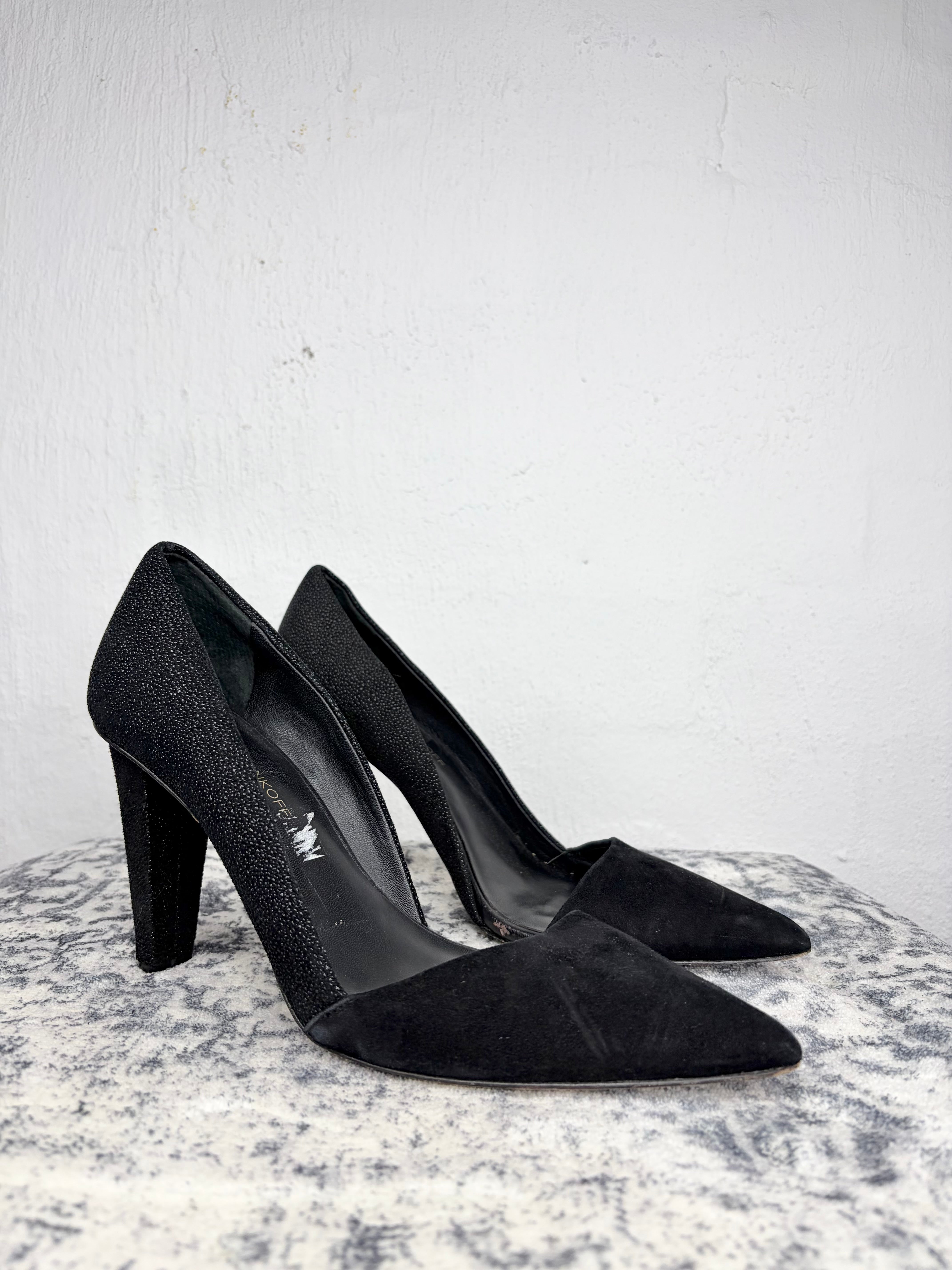 A pair of black pointed-toe high heels with a textured finish, displayed on a gray stone surface against a white wall.