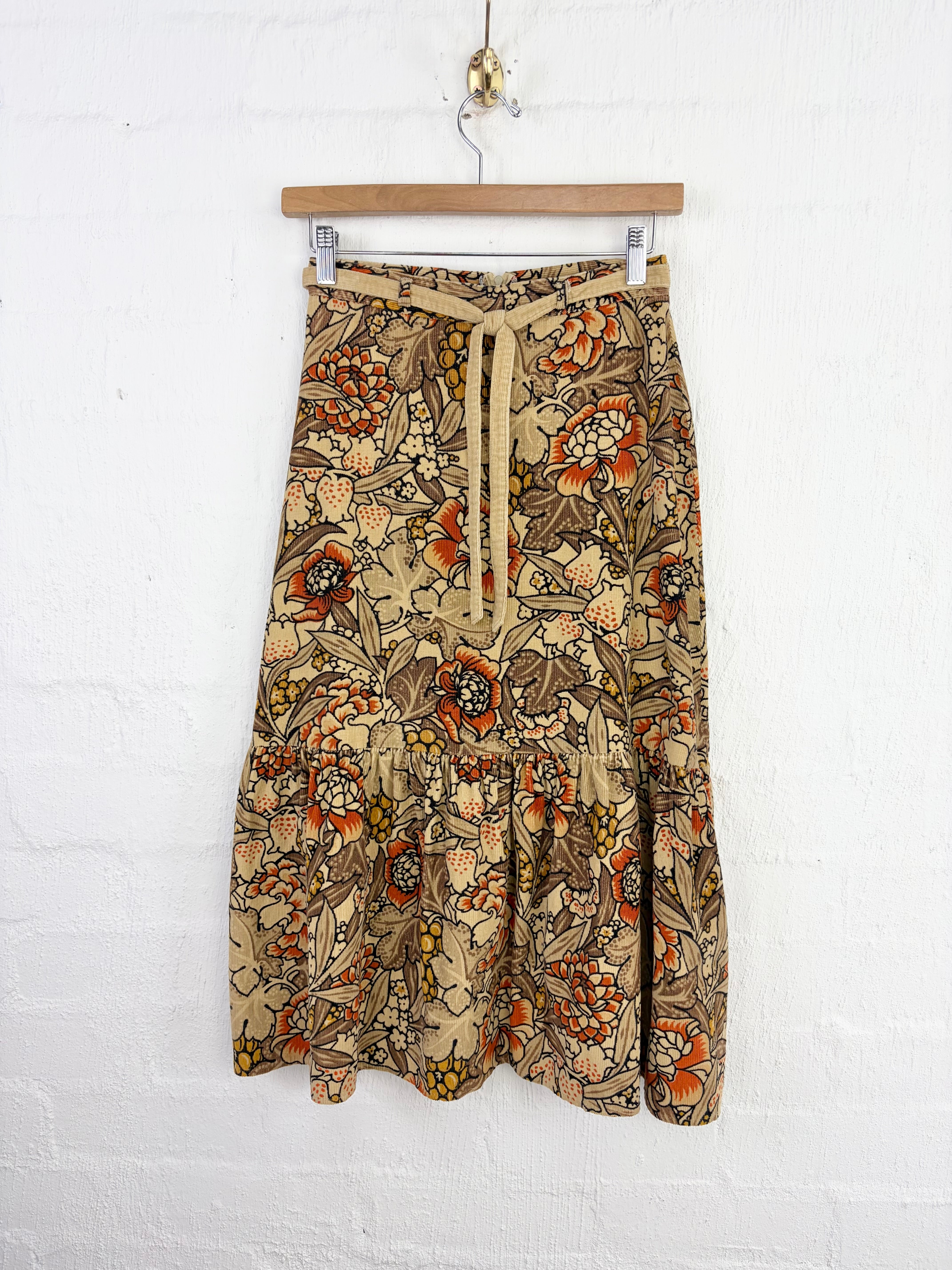 A vintage-style tiered skirt with a brown and beige floral pattern, hanging from a wooden hanger against a white painted brick wall.