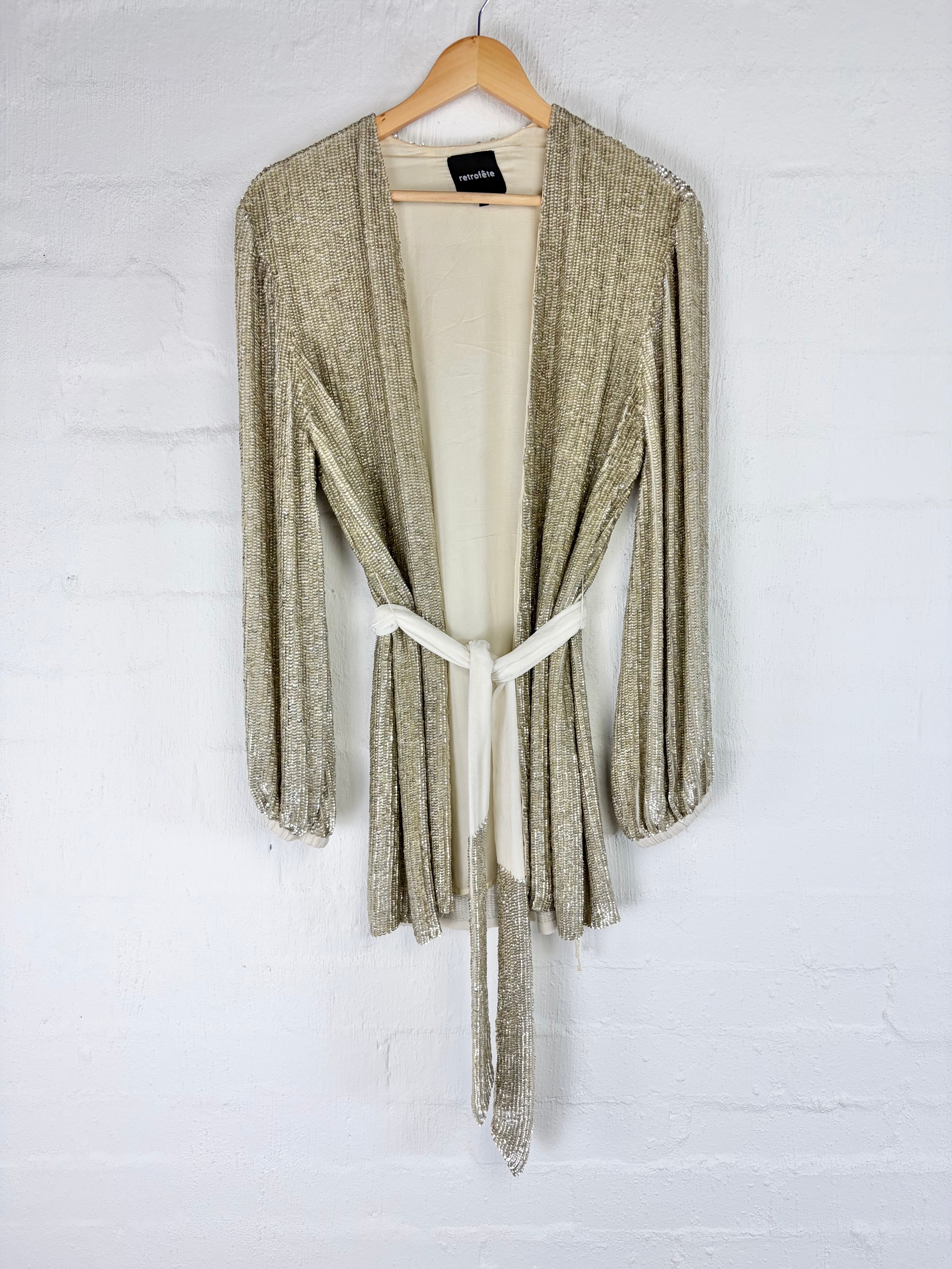 A shimmering champagne-gold open-front cardigan with long sleeves and a cream tie belt, hanging on a wooden hanger against a white painted brick wall.