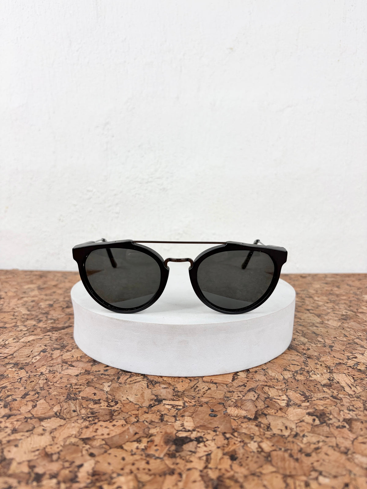 Black round-frame sunglasses with dark lenses resting on a white cylindrical pedestal over a cork surface against a white wall.
