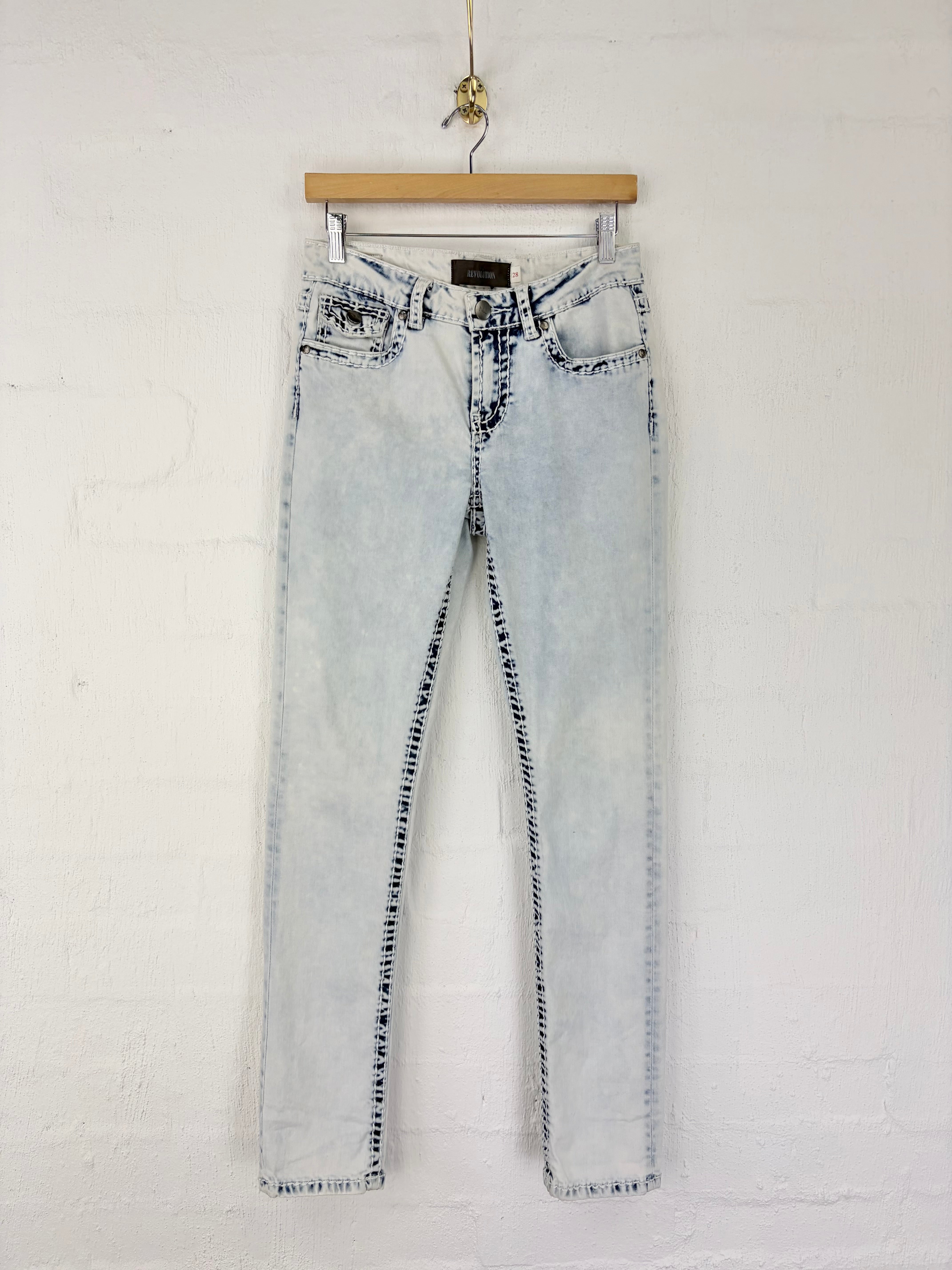 Light-wash blue denim jeans with a faded, washed-out finish and dark contrast stitching, hanging on a wooden hanger against a white painted brick wall.