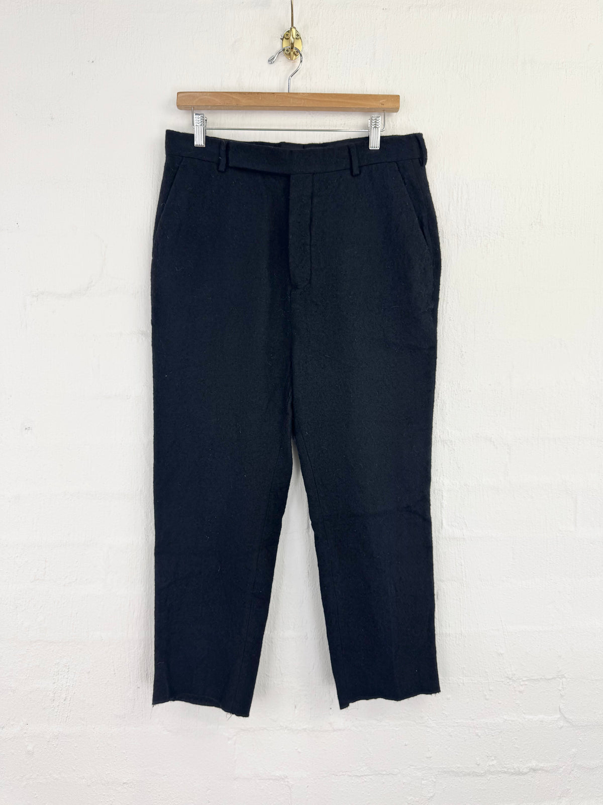 A pair of dark navy blue cropped trousers hanging from a wooden hanger against a white painted brick wall.