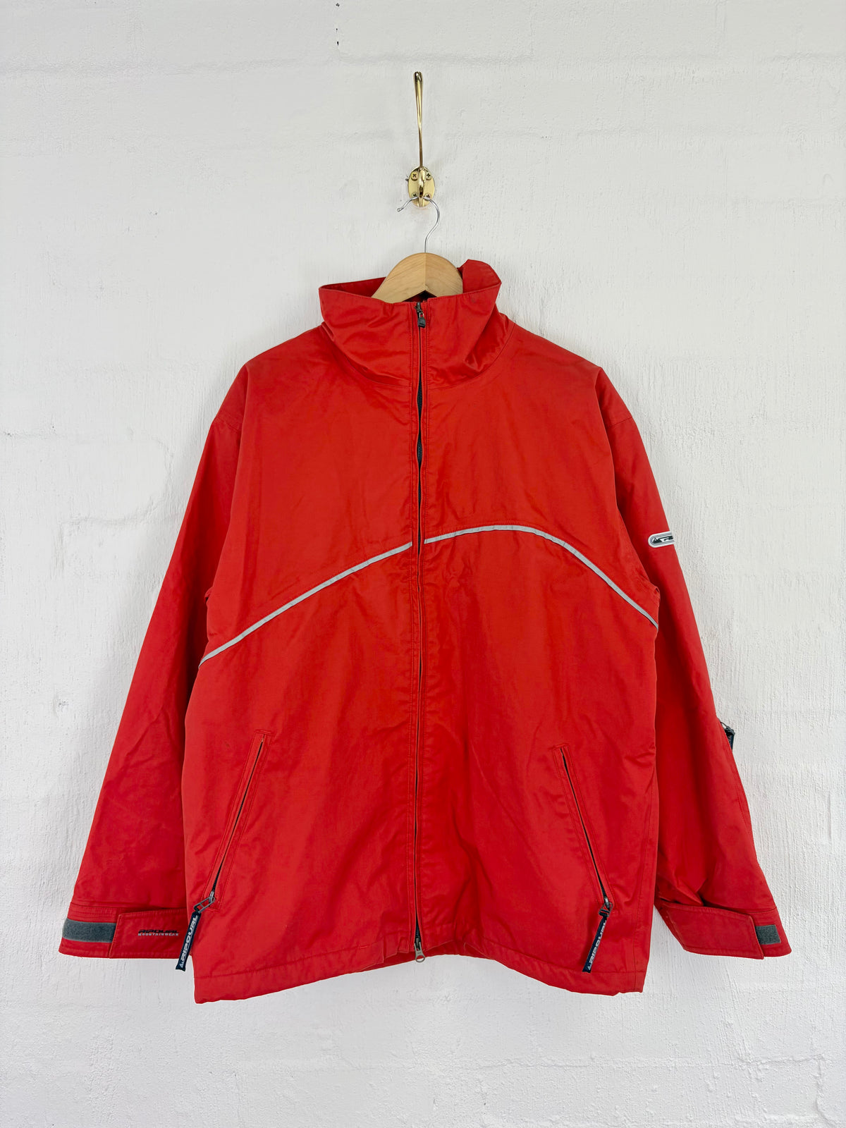 A red outdoor jacket with a high collar, front zipper, and zippered hand pockets, hanging on a wooden hanger against a white painted brick wall.