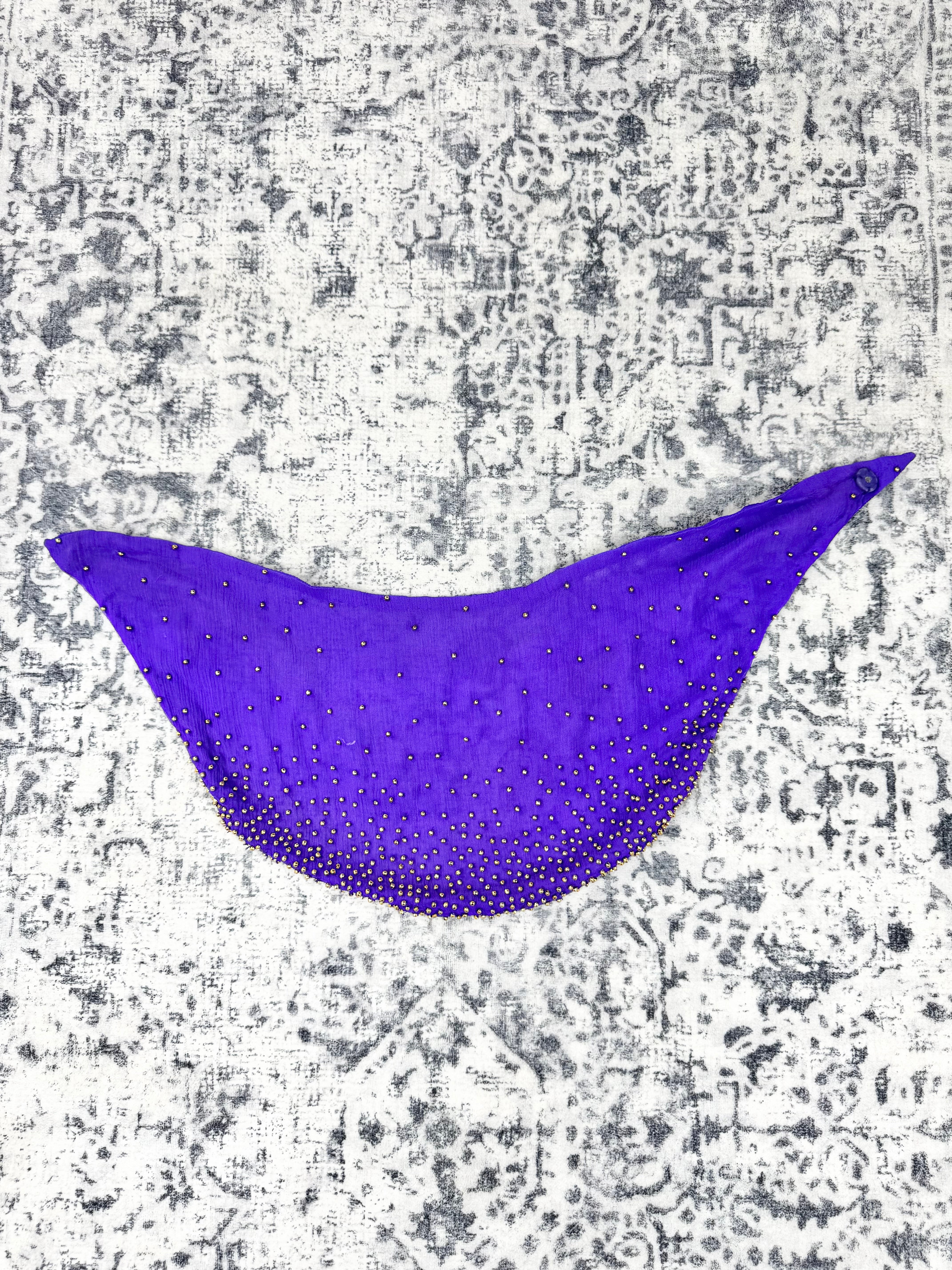 A bright purple scarf or shawl with a gradient of small gold rhinestones, laid flat against a distressed gray and white patterned background.