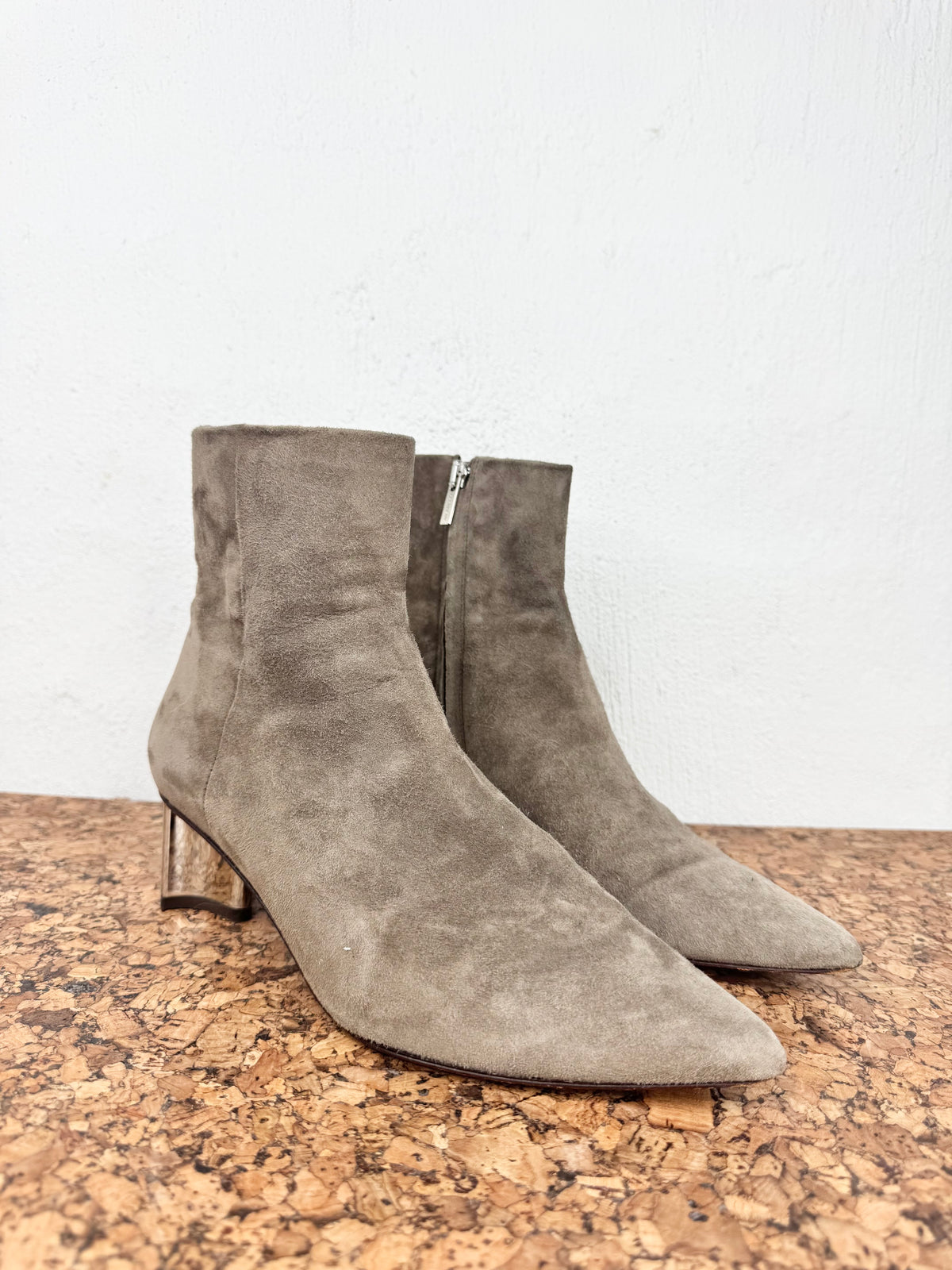 A pair of taupe-brown suede ankle boots with low block heels and pointed toes, displayed on a cork surface against a white wall.