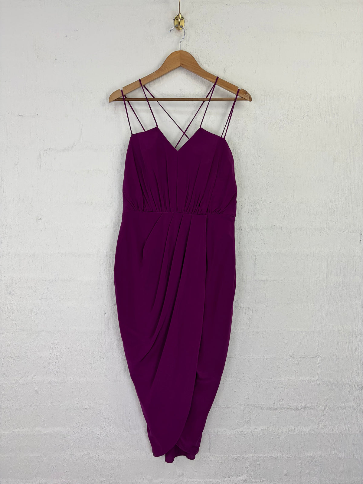 A deep purple midi-length slip dress with thin criss-cross shoulder straps, hanging on a wooden hanger against a white painted brick wall.