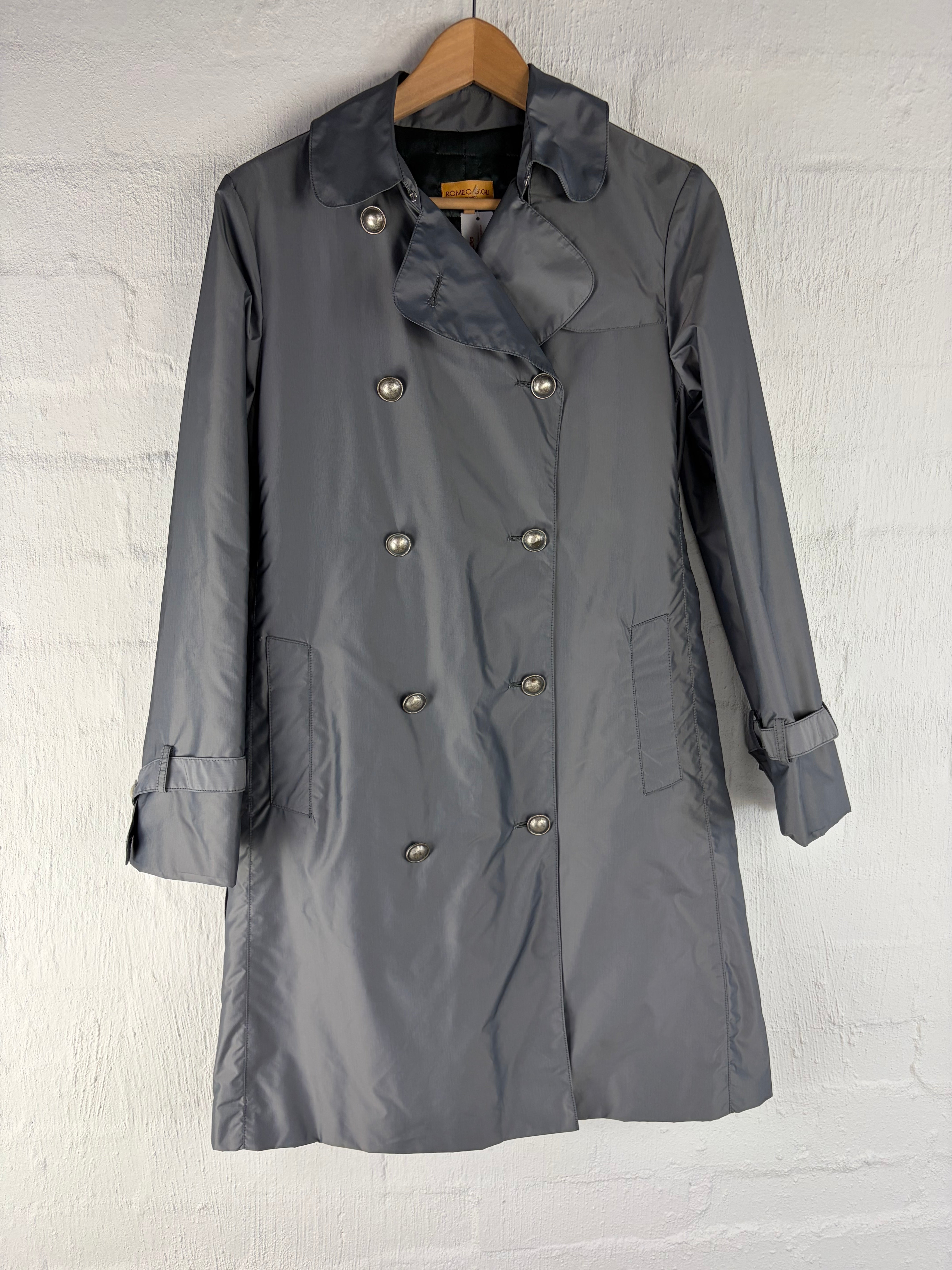 A gray double-breasted raincoat with silver buttons, laid flat on a white brick wall background.