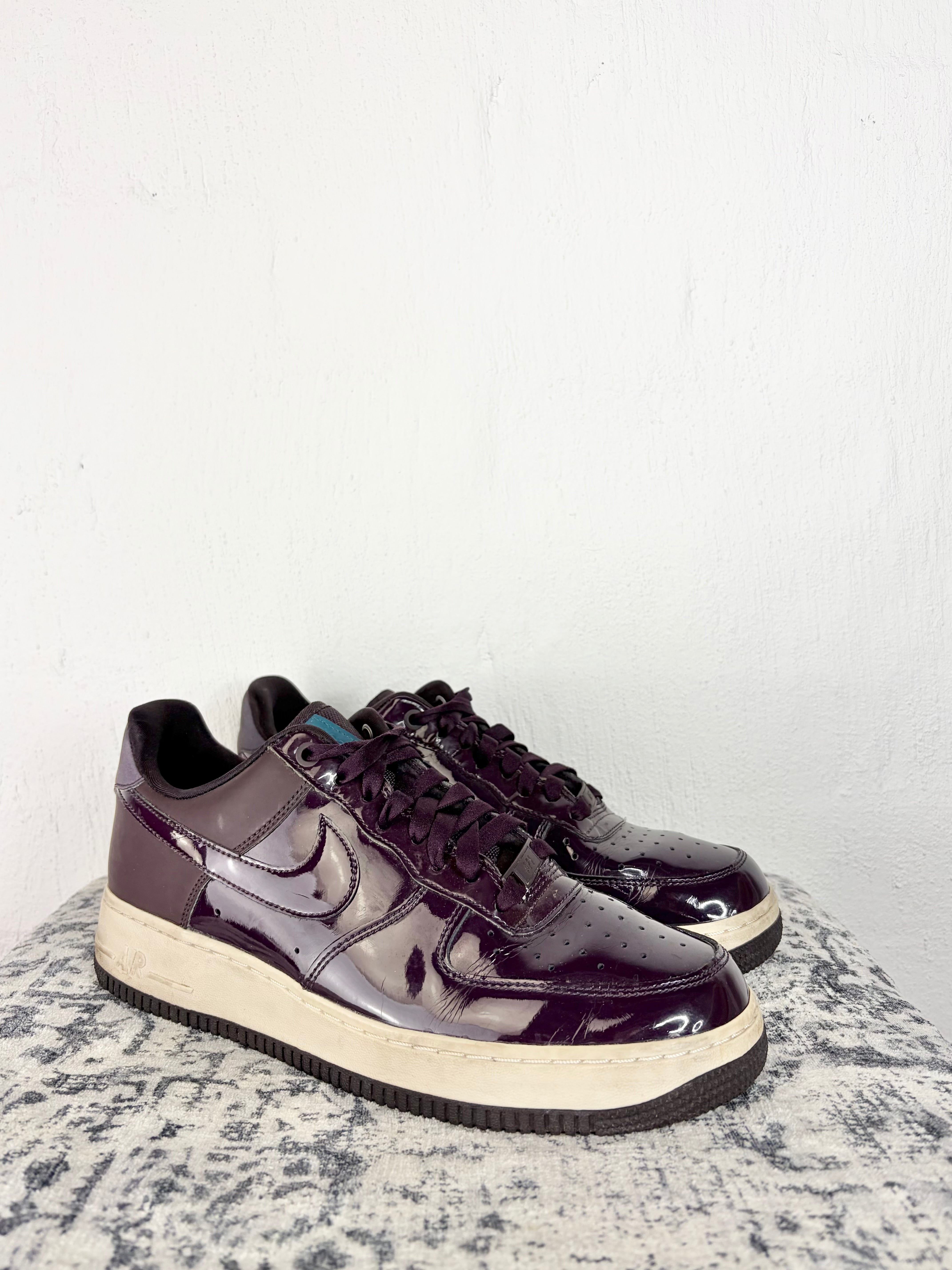 A pair of Nike Air Force 1 Low sneakers in a glossy, deep purple patent leather finish, resting on a mottled gray stone surface against a white wall.
